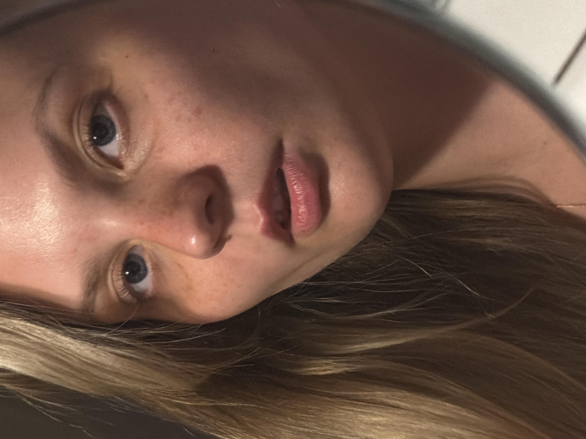 The image is a close-up, angled portrait of a young Caucasian woman with blonde hair. She has blue eyes, fair skin with freckles, and her lips are slightly parted. The lighting is warm and casts shadows on her face. The background is dark and out of focus. There is no visible indication of the city of Tel-Aviv, Israel, or any specific location within it. The image focuses solely on the woman's facial features and hair.