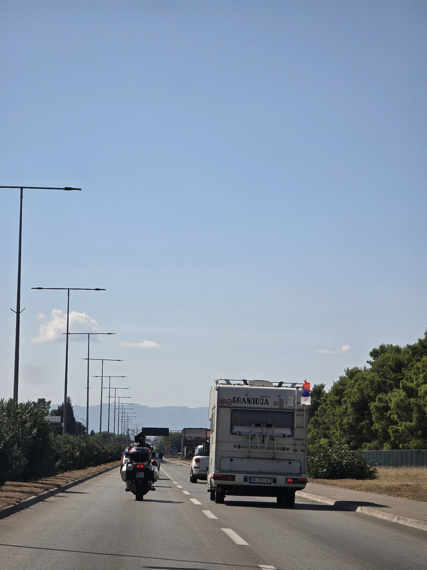 The image captures a highway scene on a clear day, likely on the outskirts of Podgorica, Montenegro, given the presence of a Serbian flag. In the foreground, a motorcycle with a rider wearing a black helmet and jacket is traveling down the road. Behind the motorcycle, a white camper van with a Serbian flag mounted on its rear is also moving. The camper van has a license plate that reads "BG 352-BT". Further ahead, a white car is visible. The road is lined with trees and streetlights, and mountains are visible in the distance. The environment appears to be semi-urban or suburban.