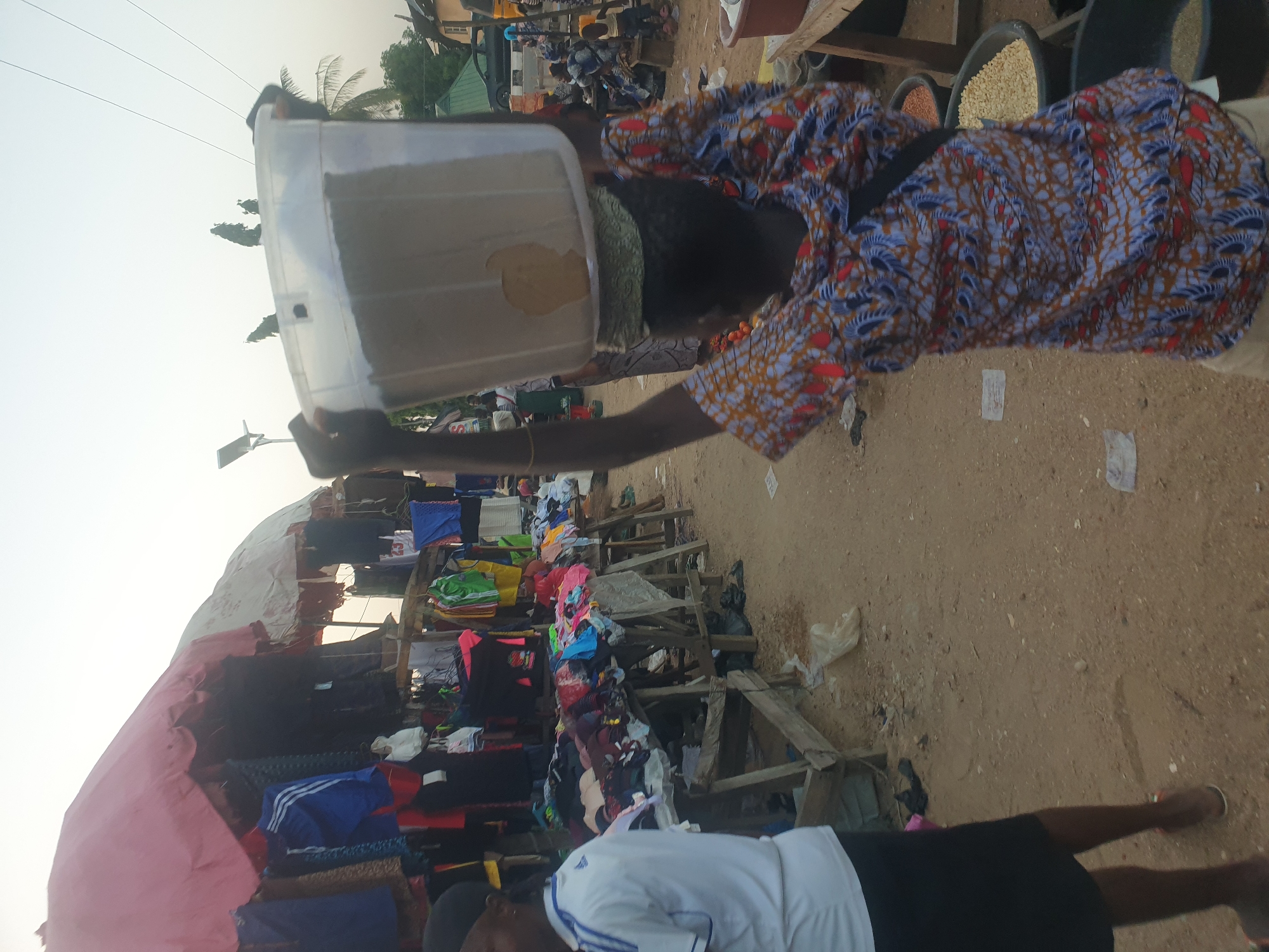 The image shows a street scene with a person in the foreground holding a clear plastic container over their head. The person is wearing a colorful patterned shirt. Behind them, there's a makeshift market stall with clothes on display under a pink awning. In the background, there are buckets filled with grains or seeds and people moving about. The ground appears to be dirt or sand. The city and location are unknown.