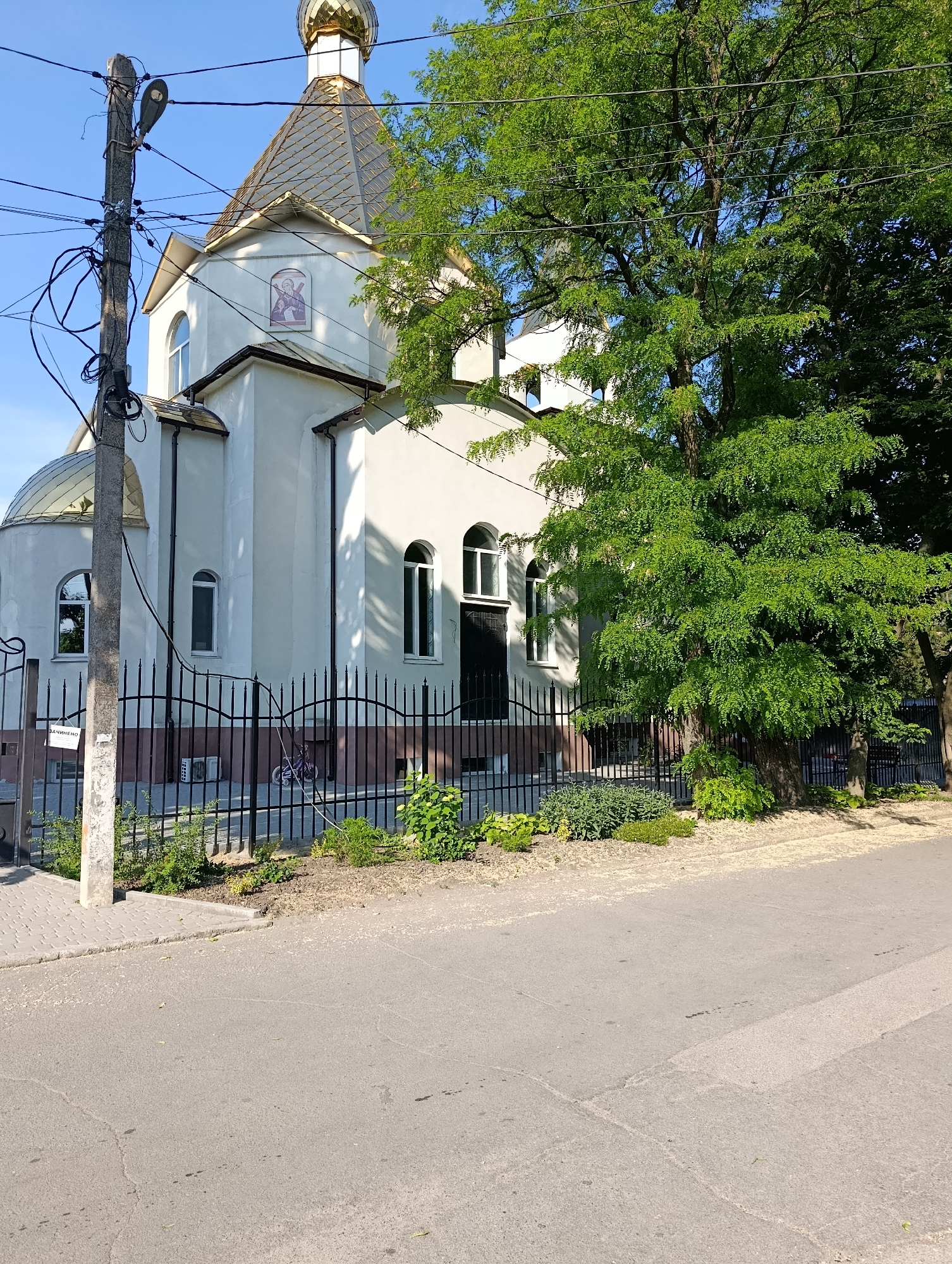A low-angle, medium shot captures the facade of a white church with a golden dome, set against a clear blue sky. The church features arched windows and a prominent icon above the entrance. A large, leafy green tree partially obscures the right side of the building. In the foreground, a grey asphalt road leads to a black metal fence surrounding the church property, behind which various plants and shrubs are visible. A utility pole cluttered with wires stands to the left of the church, and a bicycle is parked near the fence. The scene appears to be in the city of Doslidne, Ukraine.