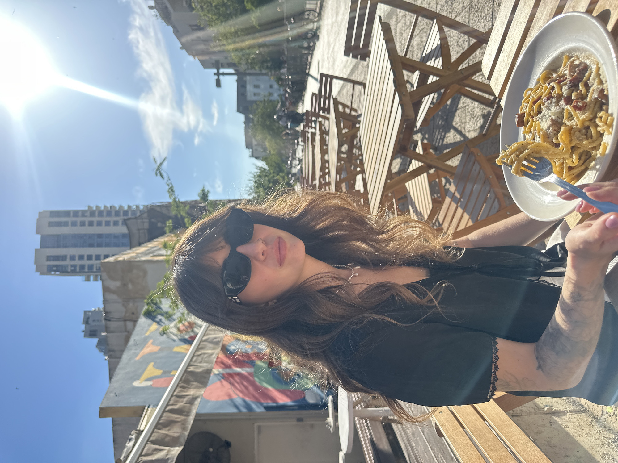 A young woman wearing sunglasses and a black blouse sits at a wooden table at an outdoor cafe in Tel Aviv, Israel. She is holding a fork with pasta on it, and a plate of pasta with bacon and parmesan cheese sits on the table in front of her. The background includes buildings, trees, and a bright sky.
