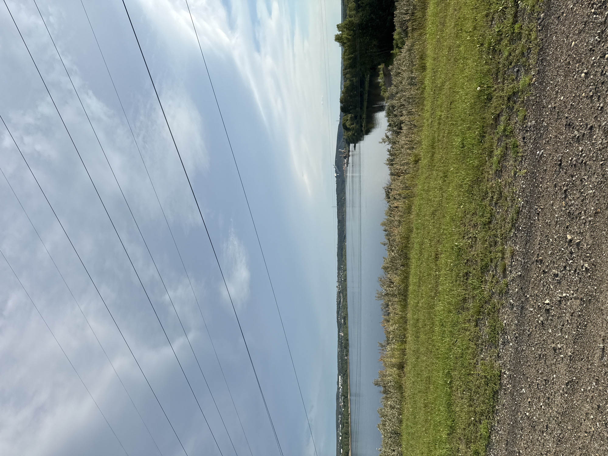 The image depicts a wide, calm body of water, likely a river or reservoir, under a partly cloudy sky. In the foreground, a grassy embankment slopes down to the water, with a gravelly path at the very bottom of the frame. Along the far bank of the water, a tree-lined shore is visible, with a town or industrial area discernible in the distance to the left. To the right, a prominent industrial structure with cranes is situated on the far bank, suggesting an active port or industrial site. Several power lines stretch diagonally across the upper portion of the image. The overall scene is serene and suggests a natural landscape near a developed area. The location is the Kama River in Chaikovsky, Russia.