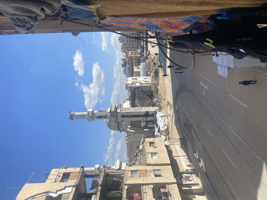 The image depicts a devastated urban street scene in Khan Yunis, Palestinian Territory, under a clear blue sky with scattered white clouds. The perspective is from an elevated vantage point, possibly a balcony, with dark clothing and a brown fabric panel visible in the immediate right foreground.
On the left, a multi-story residential building exhibits significant structural damage, with upper floors largely demolished, revealing exposed rebar and interior spaces. Laundry hangs on railings of lower-level balconies that appear less damaged. A partially visible poster is affixed to the ground floor facade.
Dominating the midground is a large mosque with a prominent, mostly intact minaret. The mosque structure itself is heavily damaged, with large sections of its roof and walls collapsed, exposing internal areas and leaving piles of rubble around its base. Debris fields extend from the damaged buildings onto the adjacent ground.
A paved asphalt road runs across the center, featuring white lane markings including a right-turn arrow. Scattered debris and patches of dirt are present along the road's edges. Several individuals are visible on the street and sidewalks: one person walks alone in dark clothing on the left side of the road, a small group of three to four individuals stands further down on the right, and other figures are dispersed in the distance. In the background, further urban buildings, some appearing damaged, and a white tent structure are visible.