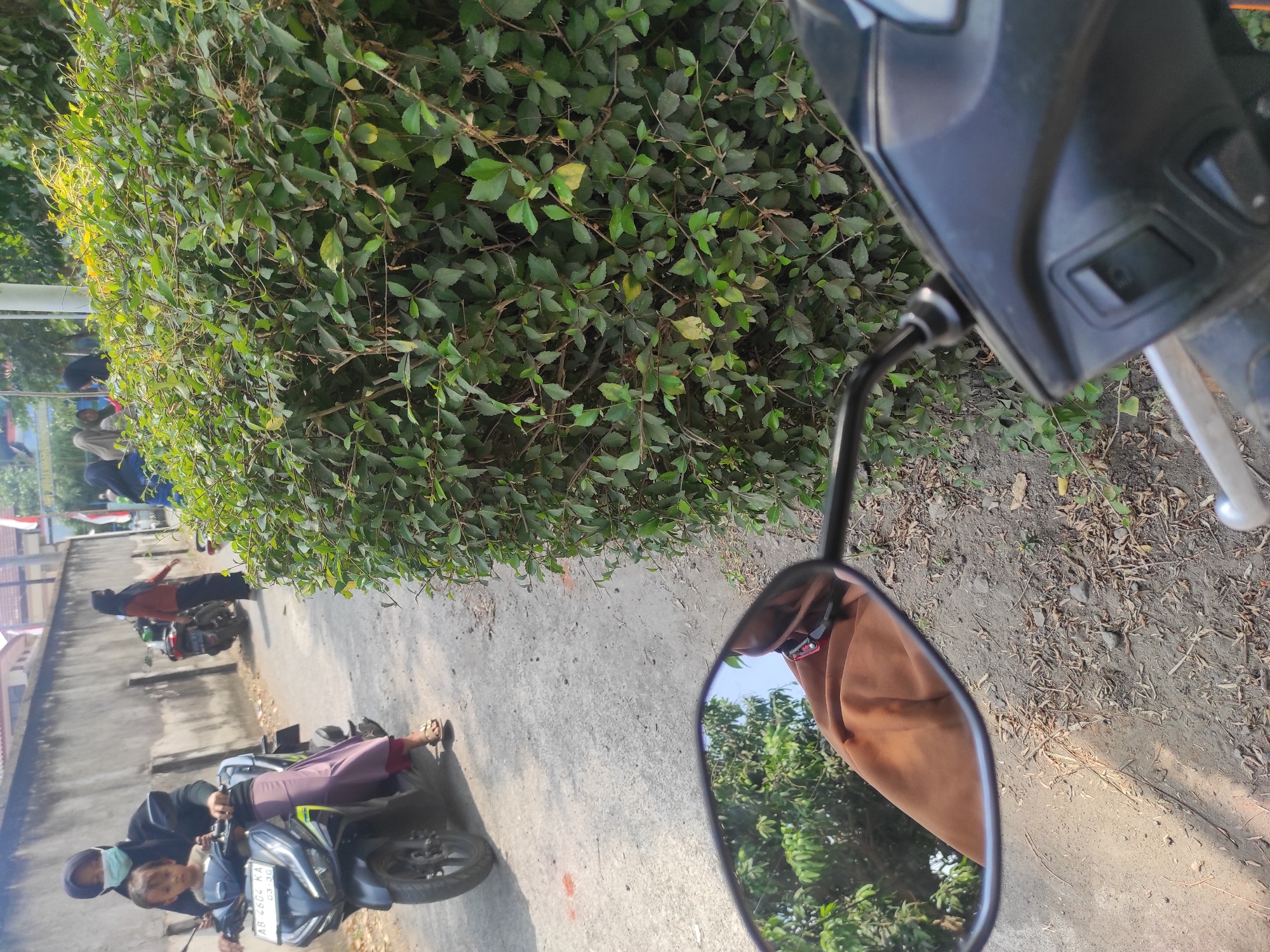 The image depicts a street scene in Pleret, Indonesia, on a sunny day. In the foreground, a motorcycle's handlebar and a side mirror are visible. The side mirror reflects trees and a person wearing a brown garment. A woman wearing a black hijab and a light blue face mask is riding a motorcycle in the middle ground, with a young child in front of her. The child is wearing a light-colored shirt. Behind them, another person in a dark hijab is walking, and further back, a person in an orange top and dark pants is also walking. A wall borders the street on the left, and a dense green bush occupies the right side of the frame. The road is paved with asphalt and appears somewhat worn, with some debris and dry leaves scattered on the side. The overall impression is a casual street scene with people on motorcycles and on foot.