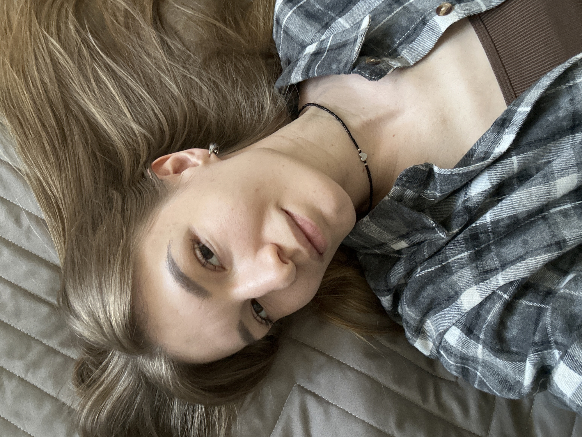 The image features a young woman captured from an overhead perspective, lying down on a textured, greyish-brown quilted surface, likely a bed or sofa. Her long, light brown hair with subtle blonde highlights is spread out around her head. She has fair skin, natural facial features with visible eyeliner, and her eyes look directly towards the viewer with a neutral, slightly pensive expression.
She is dressed in a grey and white plaid flannel shirt, which is unbuttoned to reveal a plain brown top underneath. Her accessories include a small silver hoop earring in her left ear and a delicate black beaded choker necklace adorned with a small silver heart pendant around her neck.
The activity taking place appears to be a relaxed pose or rest. The lighting is soft and even, suggesting indoor daylight conditions without harsh shadows. There are no specific environmental cues within the frame to indicate the location beyond the provided context of Chaikovsky, Russia. No text is visible in the image.