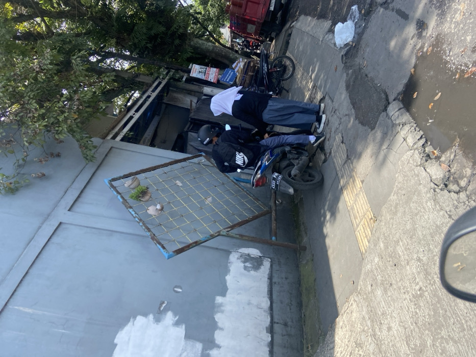 The scene is a sidewalk in the city of Kecamatan Dayeuhkolot, Indonesia. It is located at coordinates -6.963, 107.594. Two people are standing on the sidewalk. One is a man wearing a black jacket with the word "NIKE" on the back, a black helmet, and blue jeans. He is standing next to a black and blue motorcycle with a license plate that reads "4547 P 0635536". The other is a woman wearing a white hijab and a blue dress. They are both looking at each other. There is a large, metal trellis with plants growing in it, leaning against a grey building. The sidewalk is cracked and uneven, with a puddle of water in the middle. There is a green tree growing in the background, and a red truck parked on the side of the road. The truck is partially obscured by the tree and the man. There is also a black motorcycle parked on the sidewalk, behind the man. There is a white plastic bag on the ground next to the puddle.