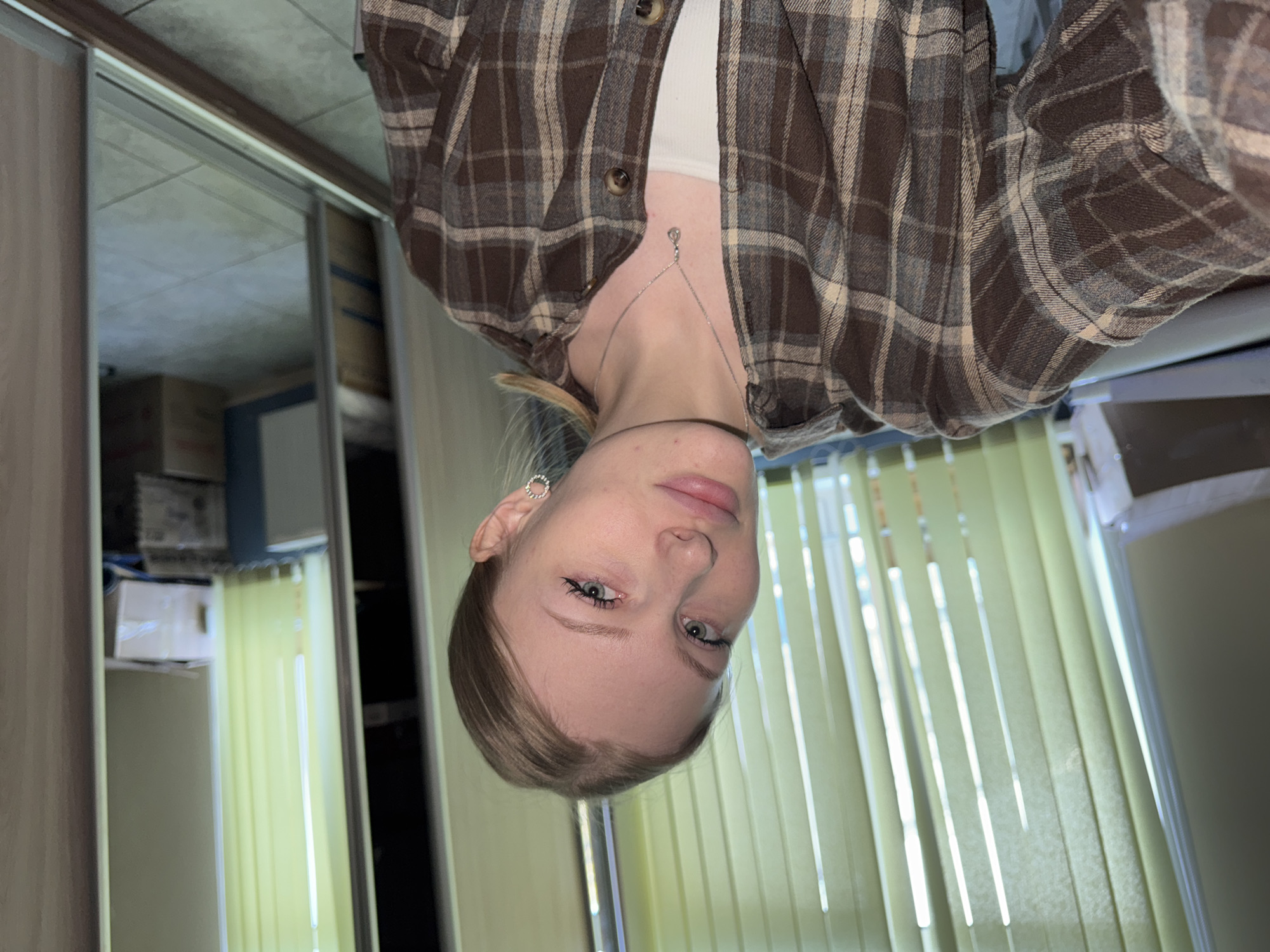 A woman with light skin and light brown hair is shown in a selfie-style photograph, slightly rotated clockwise. She wears a brown and beige plaid flannel shirt over a white top and a silver necklace. She has light blue eyes and is wearing a small, round earring. The background consists of light yellow vertical blinds and the reflection in a mirror showing storage boxes and closet contents. There is no information in the image to locate it in Perm, Russia.