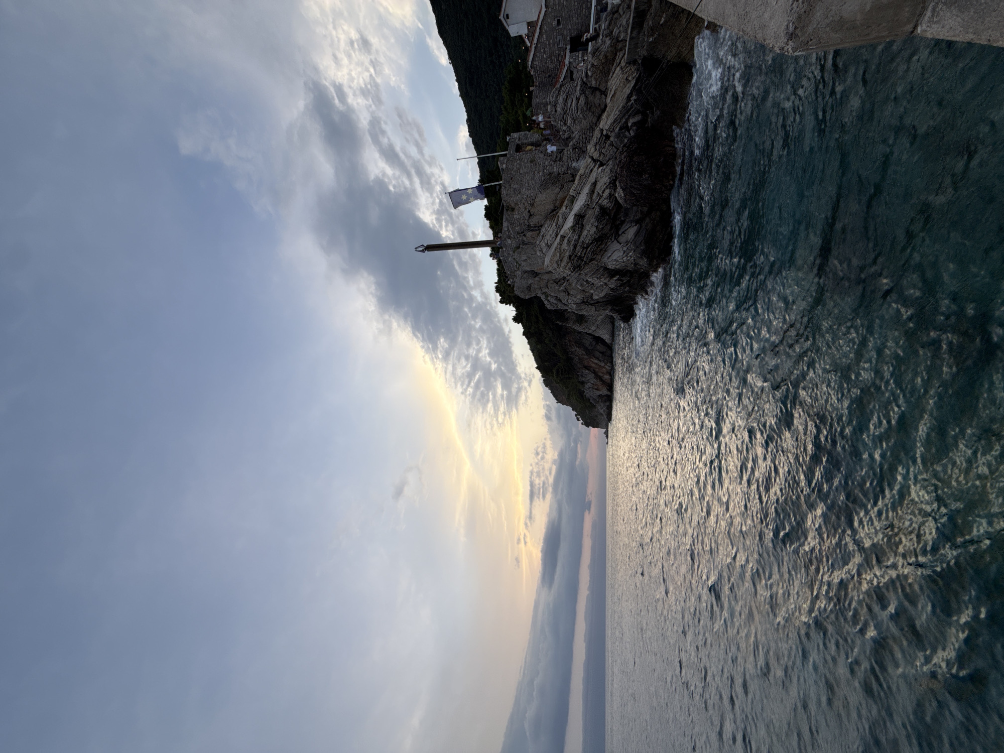 The image captures a serene twilight scene at Petrovac centar, Montenegro. The foreground is dominated by the calm, dark blue waters of the Adriatic Sea, reflecting the soft, diffused light of the setting sun. To the right, a rugged, rocky outcrop juts into the sea, crowned by ancient stone structures and lush green vegetation climbing the hillside. A tall, slender wooden structure resembling a beacon or monument stands atop the rocks. A dark blue flag with yellow stars flutters on a flagpole near the monument. Several small figures, presumably people, are visible on the stone structures, suggesting a gathering or a tourist attraction. The sky above is a gradient of soft blues and grays, with streaks of pale yellow and orange hinting at the departing sun. The overall atmosphere is peaceful and picturesque, showcasing the natural beauty and historical charm of the Montenegrin coast.