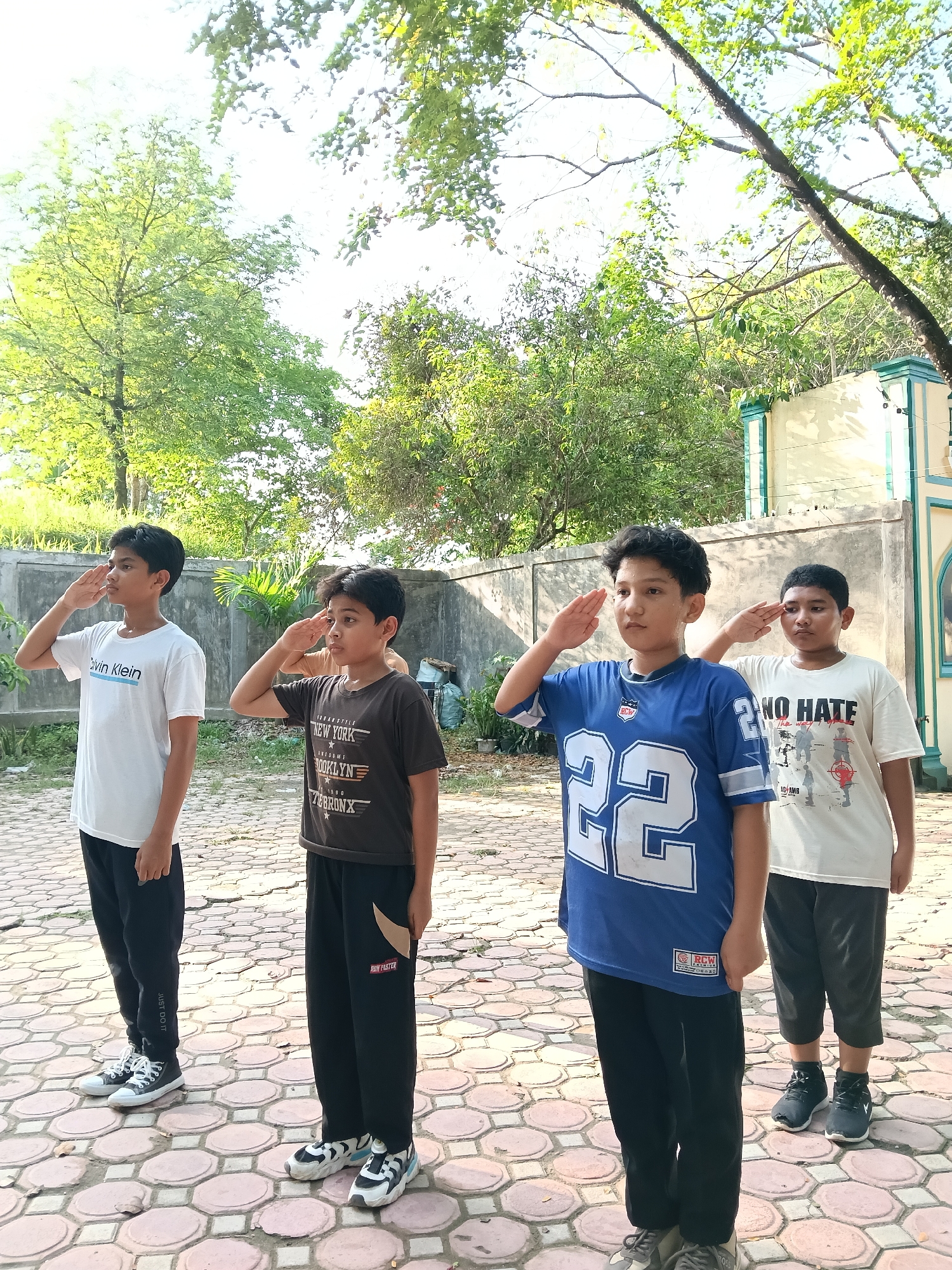 Four boys are lined up outdoors on a brick-paved surface, each saluting. The boy on the left is wearing a white t-shirt with "Calvin Klein" printed on it, black pants, and sneakers. The second boy is wearing a dark brown t-shirt with "New York Brooklyn" printed on it, black pants with "RUN FASTER" printed on them, and sneakers. The third boy is wearing a blue football jersey with the number "22" on it, black pants, and sneakers. The boy on the right is wearing a white t-shirt with "NO HATE" printed on it, dark shorts, and sneakers. In the background, there are trees, a wall, and part of a building. The location is Lhokseumawe, Indonesia.