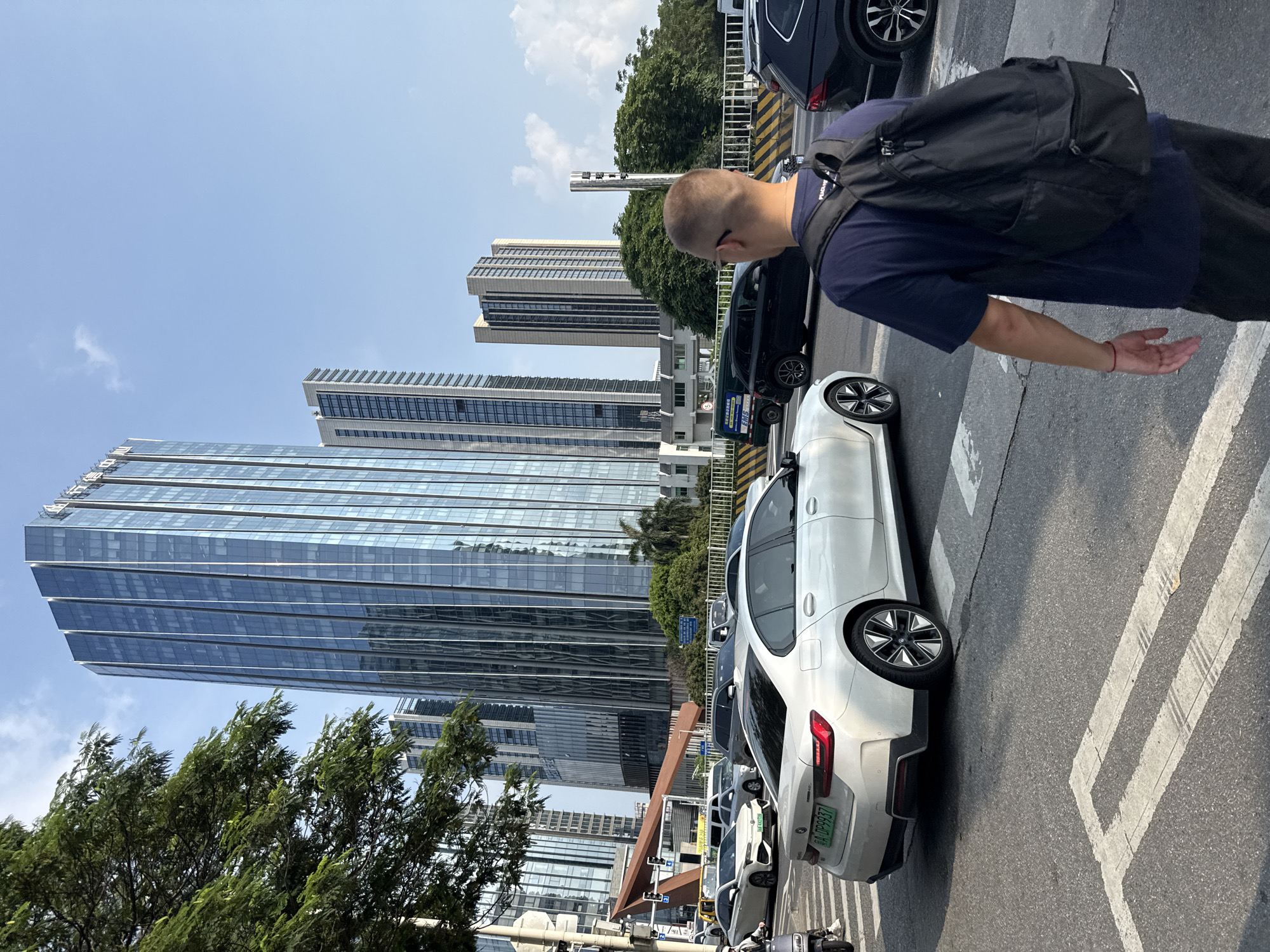 The image captures an urban street scene in Tianhe District, China, on a clear, sunny day. The primary subject is a person, seen from behind and slightly to the side, wearing a dark blue t-shirt and a black backpack. They appear to be walking across a pedestrian crosswalk or along a sidewalk adjacent to a busy street.
Several vehicles are visible on the multi-lane road, including a prominent white sedan with its rear facing the viewer, and a darker colored sedan or SUV behind it. The white sedan is positioned on or near a pedestrian crossing area.
The background is dominated by a modern cityscape featuring several tall skyscrapers with reflective glass facades, characteristic of contemporary urban development. Lush green trees are planted along the side of the road, providing a contrast to the concrete and glass. The bright daylight is evident from the strong shadows cast on the ground and the vibrant blue sky with a few faint clouds.
The activity suggests a typical urban daily commute or movement, with pedestrians and vehicles coexisting. Notable details include the architecture of the buildings, the pedestrian crosswalk markings, and the presence of traffic.
Visible text includes the license plate "A-DP9937" on the rear of the white sedan. Other text on smaller signs and vehicle license plates in the background is not clearly legible.