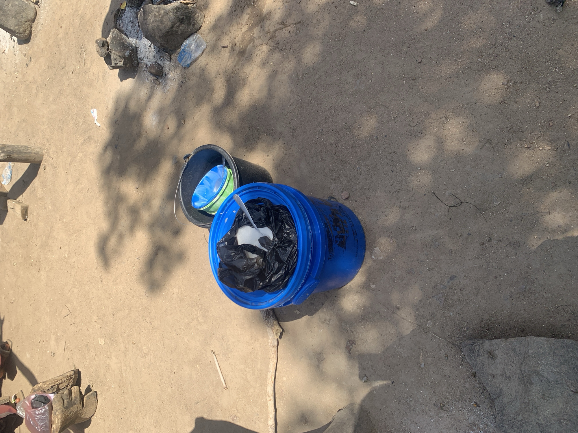 The image depicts an outdoor scene on a dirt ground, possibly in Project, Nigeria. In the foreground, a bright blue bucket is filled with black plastic bags and some white material. To the right of the blue bucket, a black bucket contains two colorful plastic bowls, one blue and one green. Scattered around the ground are rocks, a piece of wood, and some debris including plastic. Shadows from trees or structures are cast across the ground. The overall impression is one of a functional, outdoor workspace or living area, with evidence of daily activities such as the use of buckets for storage or carrying.