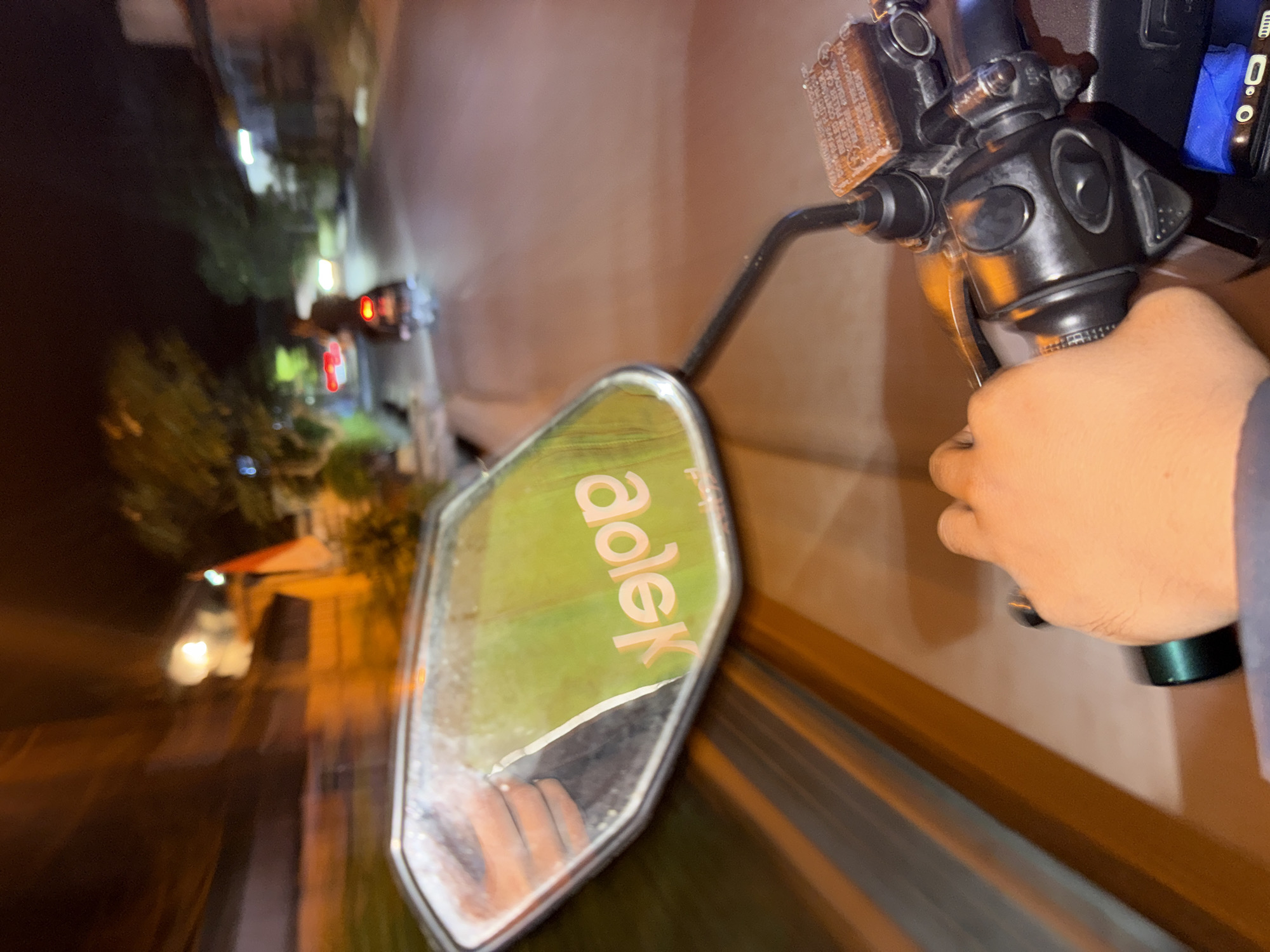 The image depicts a first-person perspective of someone riding a motorcycle at night in Denpasar, Indonesia. The scene is characterized by motion blur, suggesting the motorcycle is in motion. In the foreground, a hand is visible gripping the motorcycle's handlebar, which features various switches and a brake fluid reservoir. The rearview mirror reflects a green surface with the word "adlek" printed in white, backward, and upside down, indicating a sticker or decal. The background shows a street scene with blurred lights from passing vehicles and buildings lining the road. A flag, possibly the Indonesian flag due to its red and white colors, is visible on the right side of the frame. The overall environment suggests a typical urban street in Denpasar at night.
