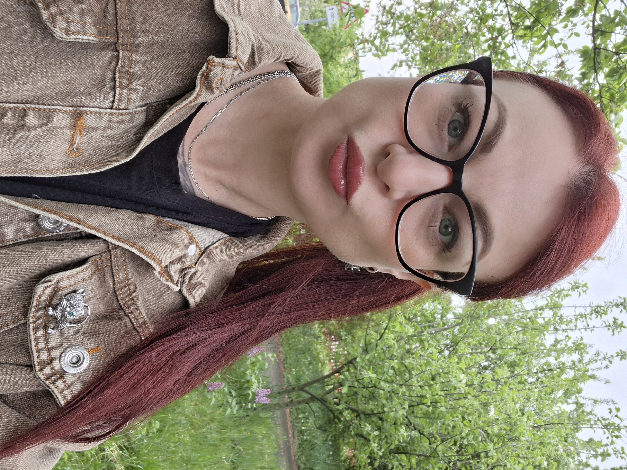 The image shows a woman with red hair wearing black-framed glasses, a black t-shirt, and a brown denim jacket with a silver cat-shaped pin. She is outdoors in Забалоцкі сельскі Савет, Belarus, as seen by the background containing trees and grass. The photo is taken at an upward angle, focusing on the woman's face.