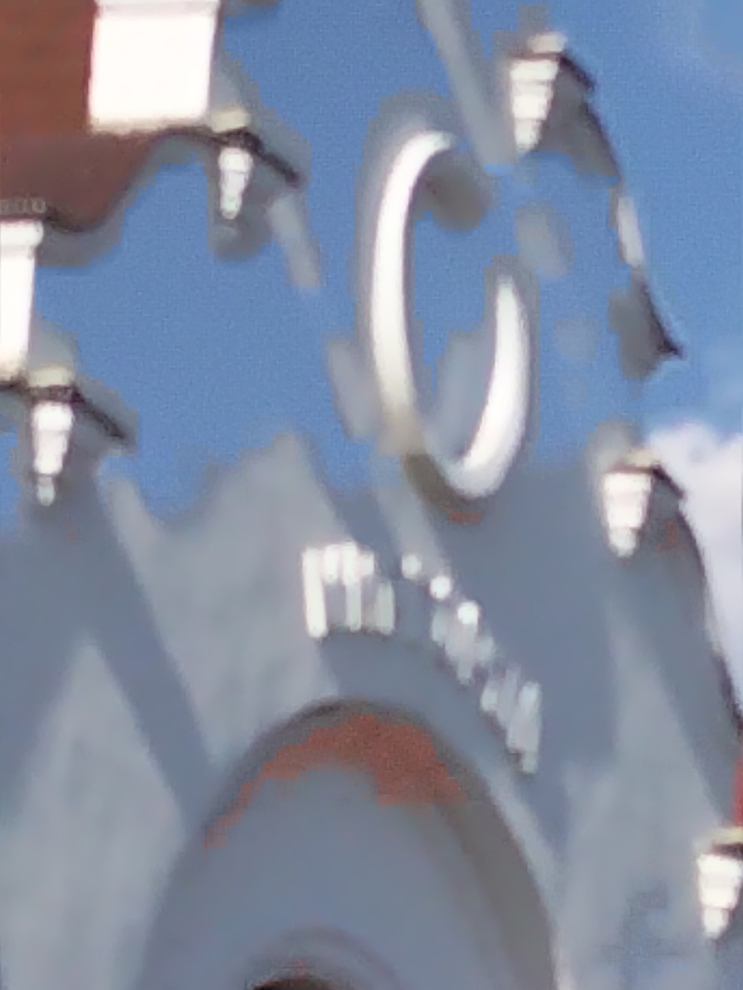 The image shows a portion of a building facade, possibly a historical structure, against a blue sky. A large white letter "C" is prominently visible, outlined in a darker color and appearing to be made of a solid material. Below the "C," there are faint, lighter-colored letters that are difficult to decipher due to the image's blurriness and resolution. These letters appear to be arranged in a curve. The building's architecture suggests it is not a modern structure, with decorative elements that are also blurred. There are no people or clear actions depicted. The overall impression is of an architectural detail of a building located in Myrhorod, Ukraine, though no specific landmarks identifiable with Myrhorod are present in the captured section. The colors are muted, with shades of blue, white, and gray dominating the scene.