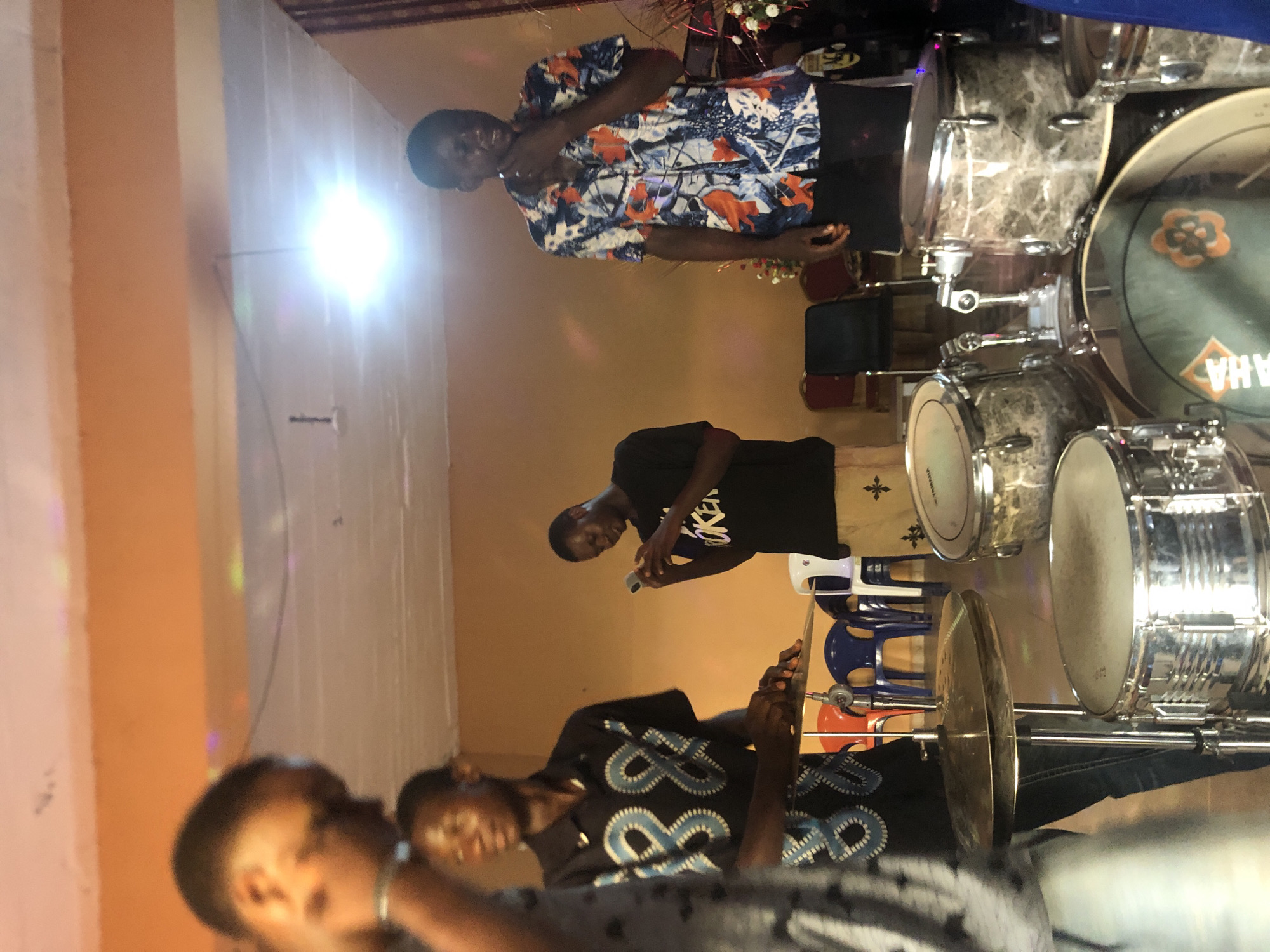 The image captures an indoor musical event or practice session, likely in Nkwelle Ezunaka, Nigeria. Three individuals are distinctly visible, with others partially obscured.
On the right side of the frame, a multi-piece drum set with a distinctive marble-like finish and chrome hardware is prominent. The bass drum head clearly displays the "YAMAHA" logo. In the upper part of the image, a man in a blue, white, and orange patterned short-sleeved shirt stands, looking towards the viewer with a slight smile, his hand near his mouth as if singing or speaking into a microphone.
In the mid-ground, a man wearing a black t-shirt with the white text "BROKEN" is looking down at a mobile phone he holds in his hands. To the lower left, a person is seated behind a cymbal, wearing a dark shirt with a white and light blue patterned design, appearing to play the drums. Another person is partially visible on the far left, out of focus.
The setting is an indoor space with light-colored walls and some ceiling beams. A very bright light source on the left side of the room creates a strong glare. In the background, blue and red plastic chairs are visible, along with some floral decorations. The atmosphere suggests a focused musical activity.
