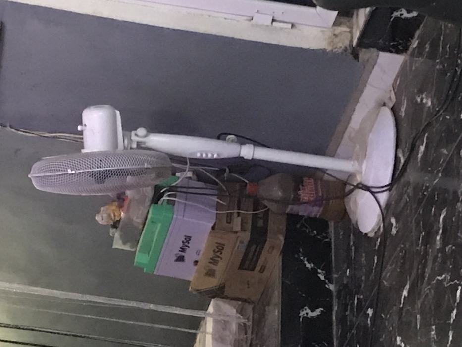 A white standing fan with a cage leans towards the left, partially obscuring a messy collection of items. These items include a green-lidded MySol battery box, a cardboard box with "MySol" printed on it, a plastic bottle with a red cap, and miscellaneous items wrapped in plastic. The background features a gray wall, a white window frame, and a marble-patterned floor. Electrical wires snake behind the fan.