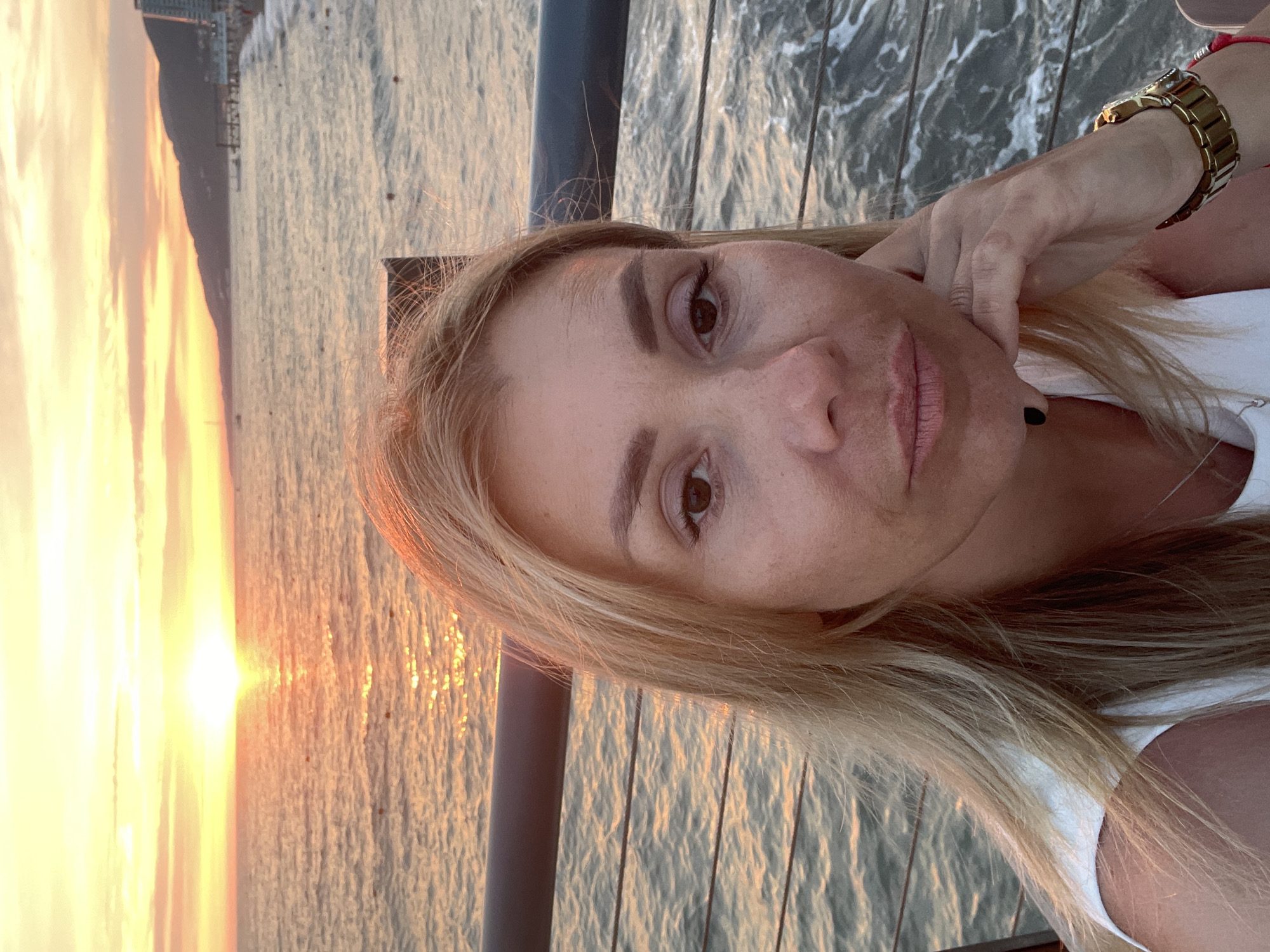 The media file contains a selfie taken by a woman with blonde hair. She is wearing a white top and a gold watch on her left wrist. Her right hand is resting on her chin. The background shows a body of water reflecting the warm glow of a sunset. A dark railing runs along the bottom of the frame. The overall lighting suggests it is sunset or sunrise. The woman's expression appears relaxed and contemplative. No text is visible in the image.