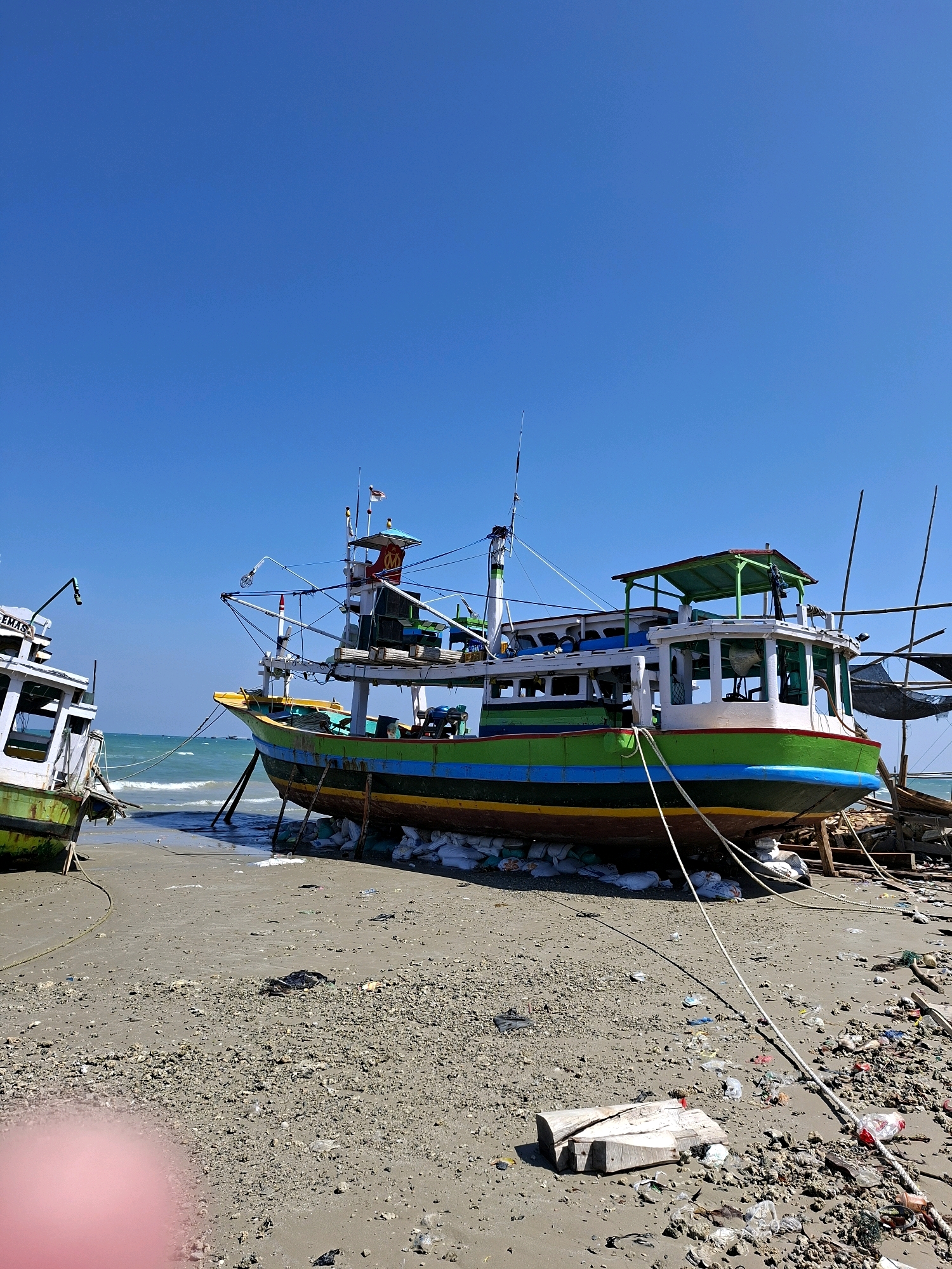 A low-angle, full shot captures a bright, sunny day at the Camplong beach in Camplong, Indonesia. The scene is dominated by two fishing boats grounded on the sandy shore. The larger boat, in the foreground, is brightly painted with horizontal stripes of green, blue, and yellow, resting on a bed of white sandbags. The smaller boat, to the left, is partially visible and appears to be white with green trim. The ocean stretches out behind the boats, with gentle waves lapping the shore under a clear blue sky. The beach itself is littered with debris, including plastic bottles, bags, and pieces of wood, indicating a potential environmental concern. There are no people visible in the image.