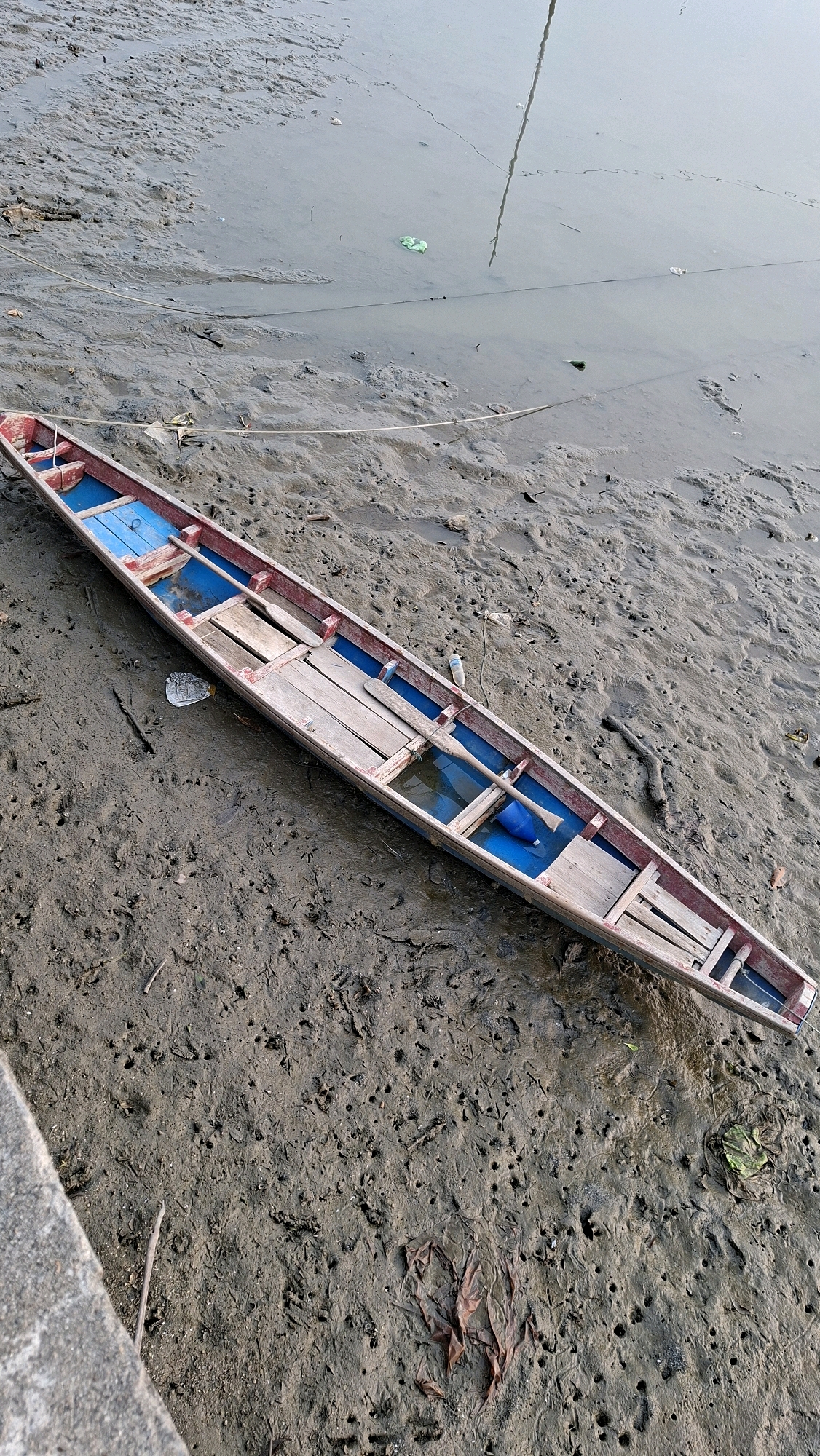 The image shows a long, narrow wooden boat with a red trim resting on muddy ground next to a body of water. The boat has blue flooring and several wooden planks forming a seating area. Two paddles are visible inside the boat, and a blue plastic container is also present. The muddy ground is uneven and shows signs of shallow water or recent flooding, with small holes and scattered debris, including leaves and what appears to be a discarded plastic bottle. The water in the background is calm and appears murky. The scene is likely a riverside or coastal area in Lhokseumawe, Indonesia, given the type of boat and the natural environment depicted.