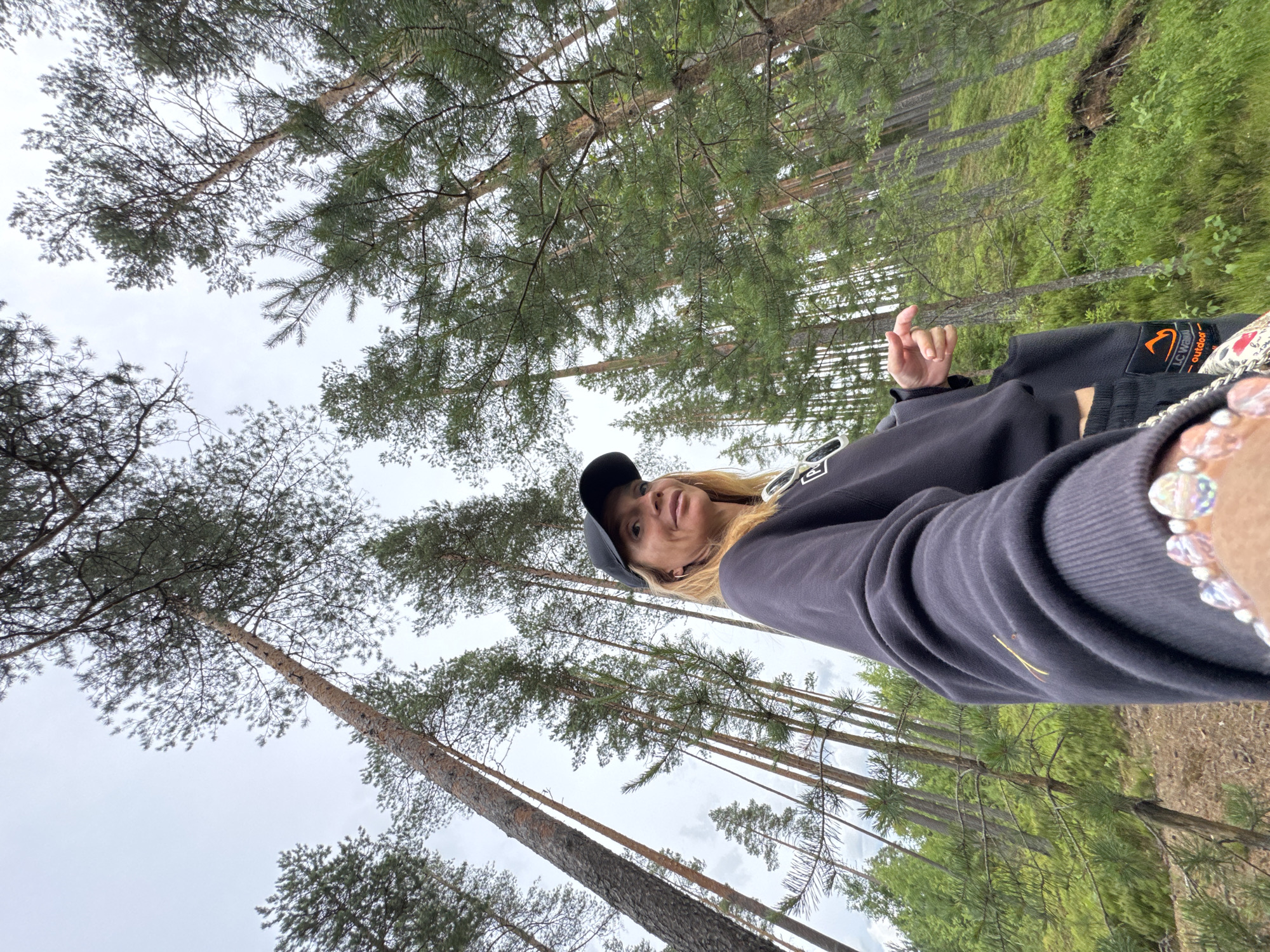A low-angle, wide shot captures a woman with blonde hair and fair skin standing in a pine forest. She is wearing a black baseball cap, a dark grey sweatshirt, and dark pants with an orange logo on the right leg. Her left arm is extended towards the camera, and her left hand is visible, adorned with a bracelet of colorful beads. She is looking slightly to her right, with her mouth open. The background is dominated by tall, slender pine trees with rough bark, reaching towards a cloudy sky. The forest floor is covered in green undergrowth and pine needles. The scene appears to be in the city of Peski, Russia.