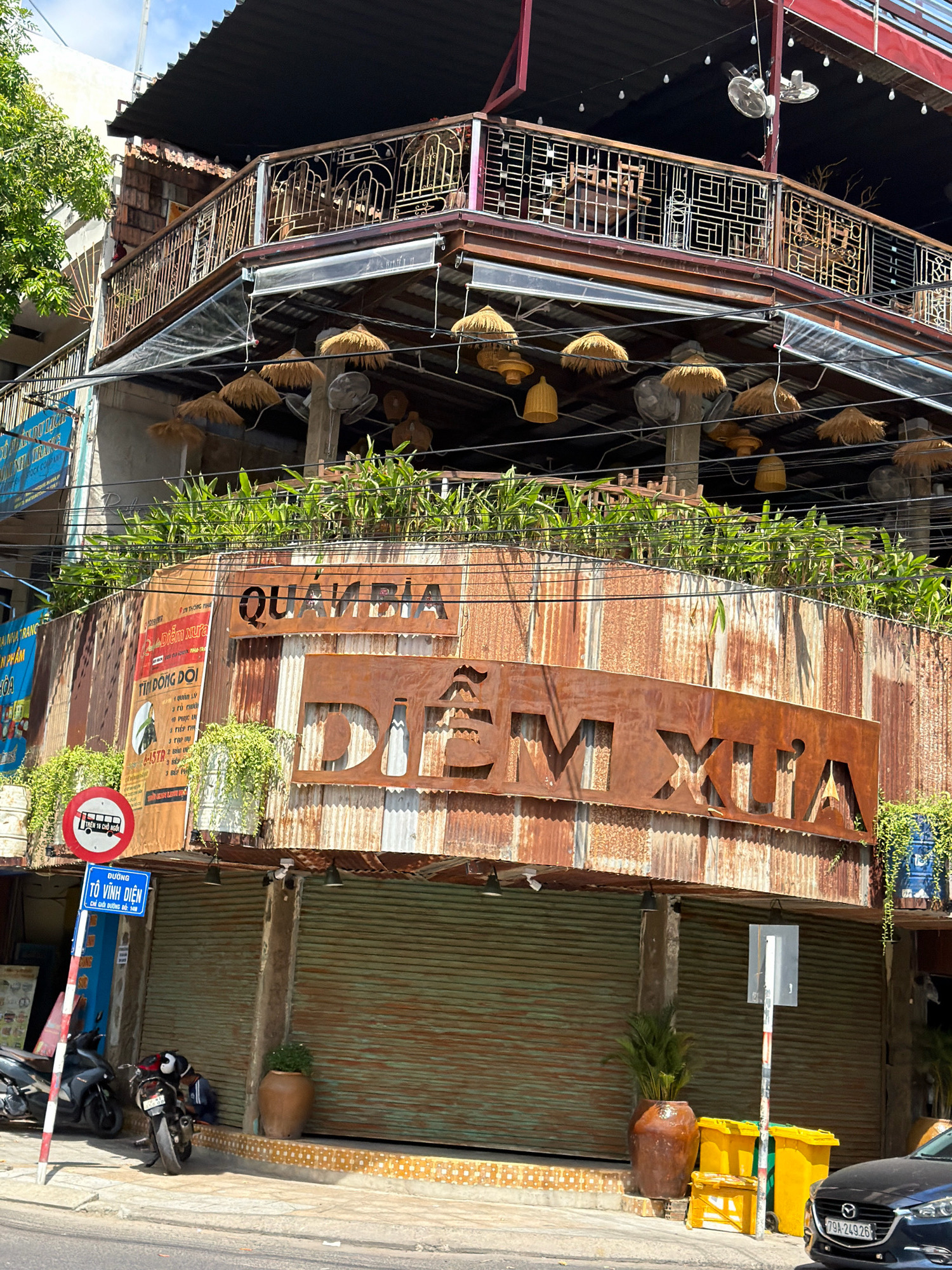 The image captures a street scene in Nha Trang, Vietnam, featuring a multi-level building with a rustic, weathered facade. The ground floor is occupied by shuttered storefronts, one of which has a green metal roller door. Above the storefronts, the building's facade is constructed from corrugated, rusted metal, giving it an aged appearance.
Prominently displayed on this rusted surface are large, cut-out metal signs. The upper sign reads "QUÁN BIA" (Beer Garden) in a stylized font. Below this, a larger sign spells out "DIEM XUA" (Old Place) in even bolder, rustic lettering. This sign has an image of a beer bottle integrated into the "X" of "XUA".
Above the rusted facade, the building features a balcony with decorative metal railings. Hanging lights with woven straw shades and several ceiling fans are visible, suggesting an outdoor seating area. Lush green plants spill over the edge of the building above the rusted facade, adding a touch of greenery to the urban environment.
On the sidewalk in front of the building, several objects are present. A red and white striped pole supports two signs: a circular red sign with a white border depicting a bus crossed out, indicating no buses, and a blue rectangular sign with Vietnamese text, likely street or district information. Motorcycles are parked on the sidewalk. Two large yellow trash bins and a smaller yellow container are situated near the building's entrance. A dark-colored car with a Vietnamese license plate (79A-249.26) is parked on the street.
A person, wearing a dark blue shirt and light-colored pants, is seated on the ground next to a motorcycle. The overall impression is a blend of commercial activity and urban decay, with a focus on a retro or vintage aesthetic for the establishment. The lighting suggests it is daytime.