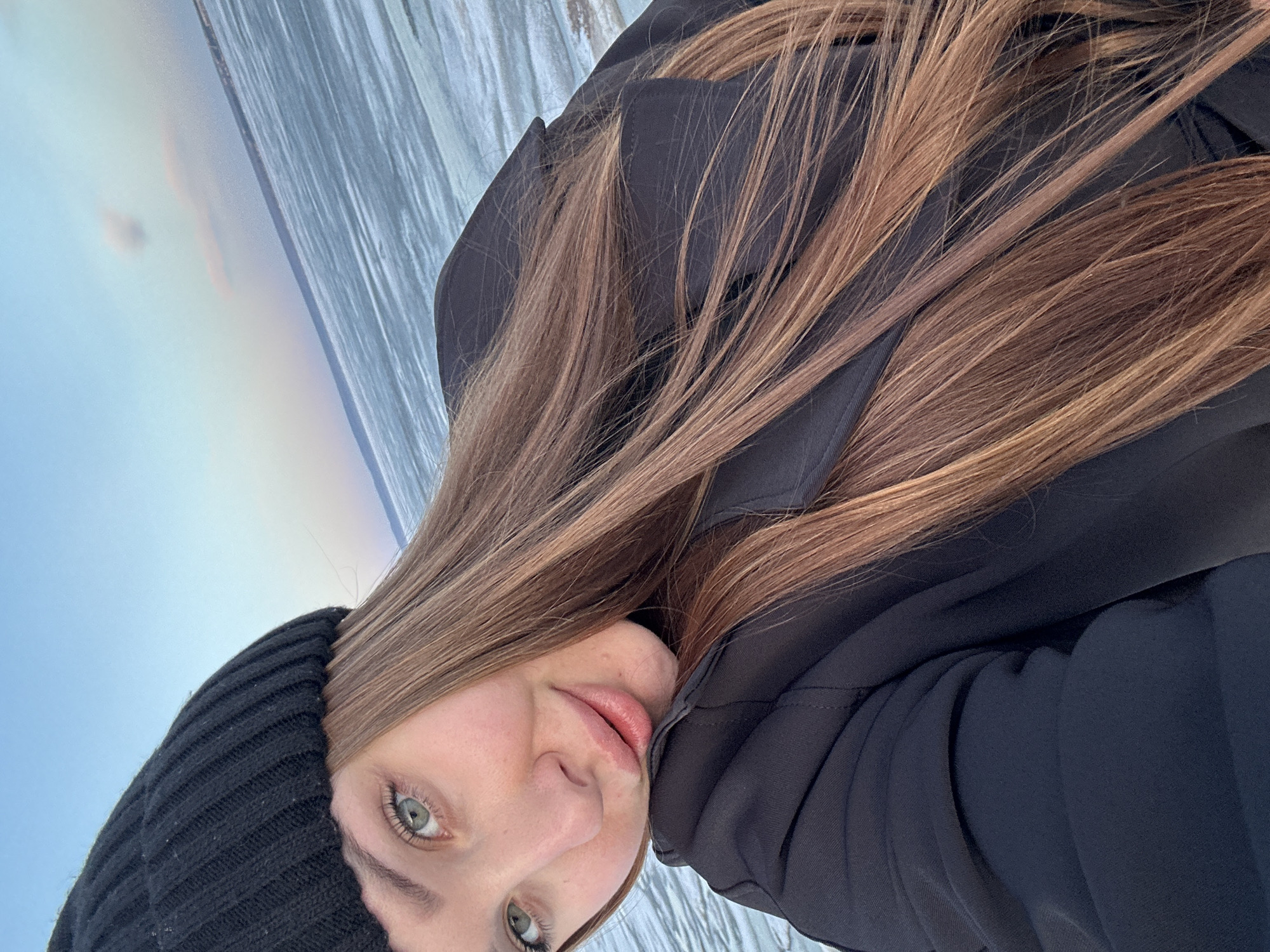 A young woman in a black knitted hat and a black jacket stands outside. In the background is a frozen river with snow cover. A faint rainbow-like light effect is visible in the sky near the river. The location is inferred to be Chaikovsky, Russia, based on the prompt.