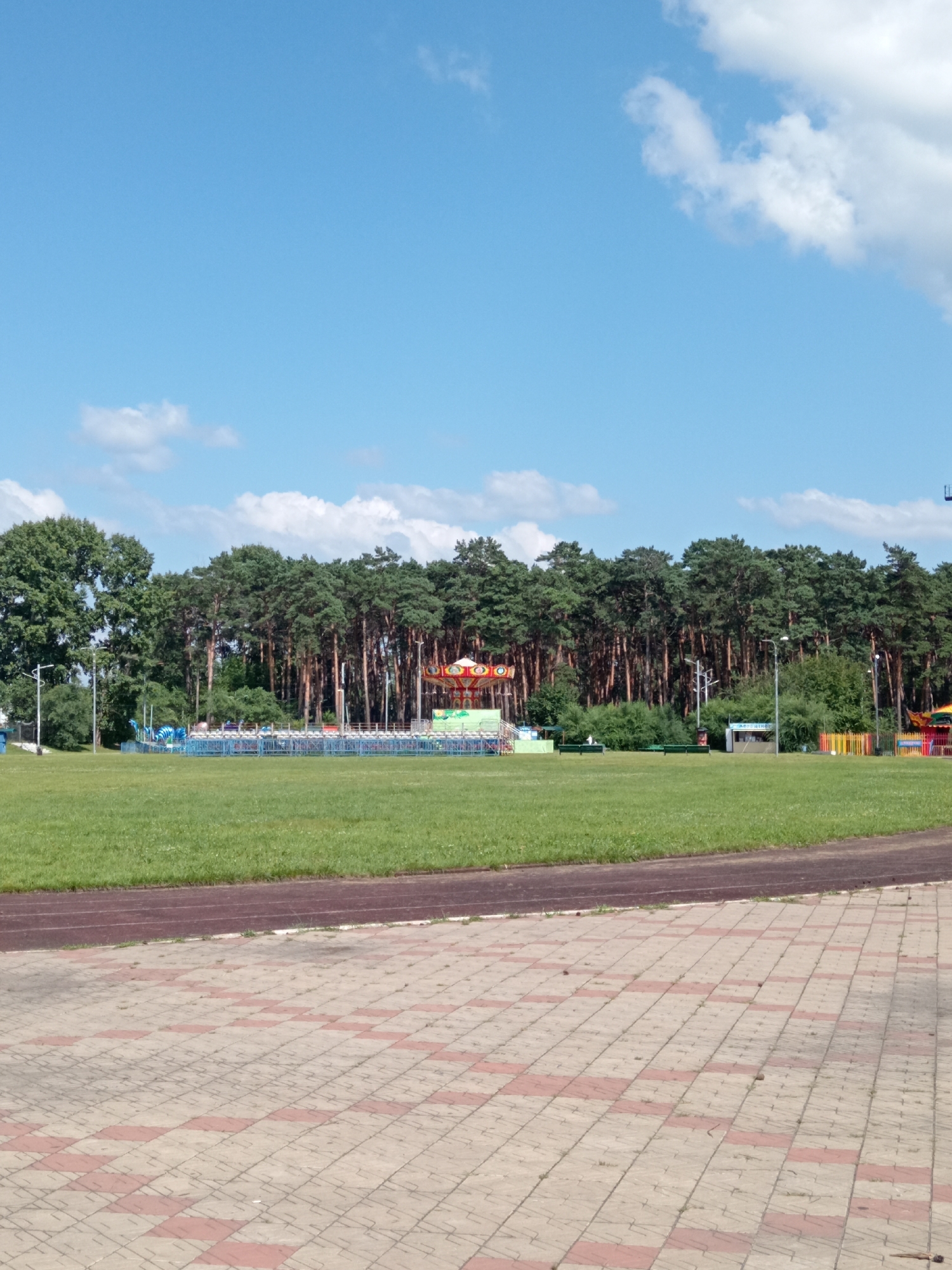 The image depicts an outdoor scene in Belogorsk, Russia, likely a public park or recreational area. In the foreground, a paved walkway with a red and beige diamond pattern leads the eye towards a vast expanse of green grass, suggesting a sports field or parkland. A dark reddish-brown running track borders the grassy area. In the midground, a row of tall, mature pine trees forms a dense backdrop. Scattered amongst the trees and the edge of the field are several amusement park rides and structures, including a carousel with red and gold accents, a blue dinosaur-themed ride, and a smaller, colorful ride with yellow and red fencing. A small blue structure and a green booth are also visible. The sky above is a bright blue with scattered white clouds. No people are clearly visible in the frame. The overall impression is one of a sunny day at a family-friendly outdoor venue.