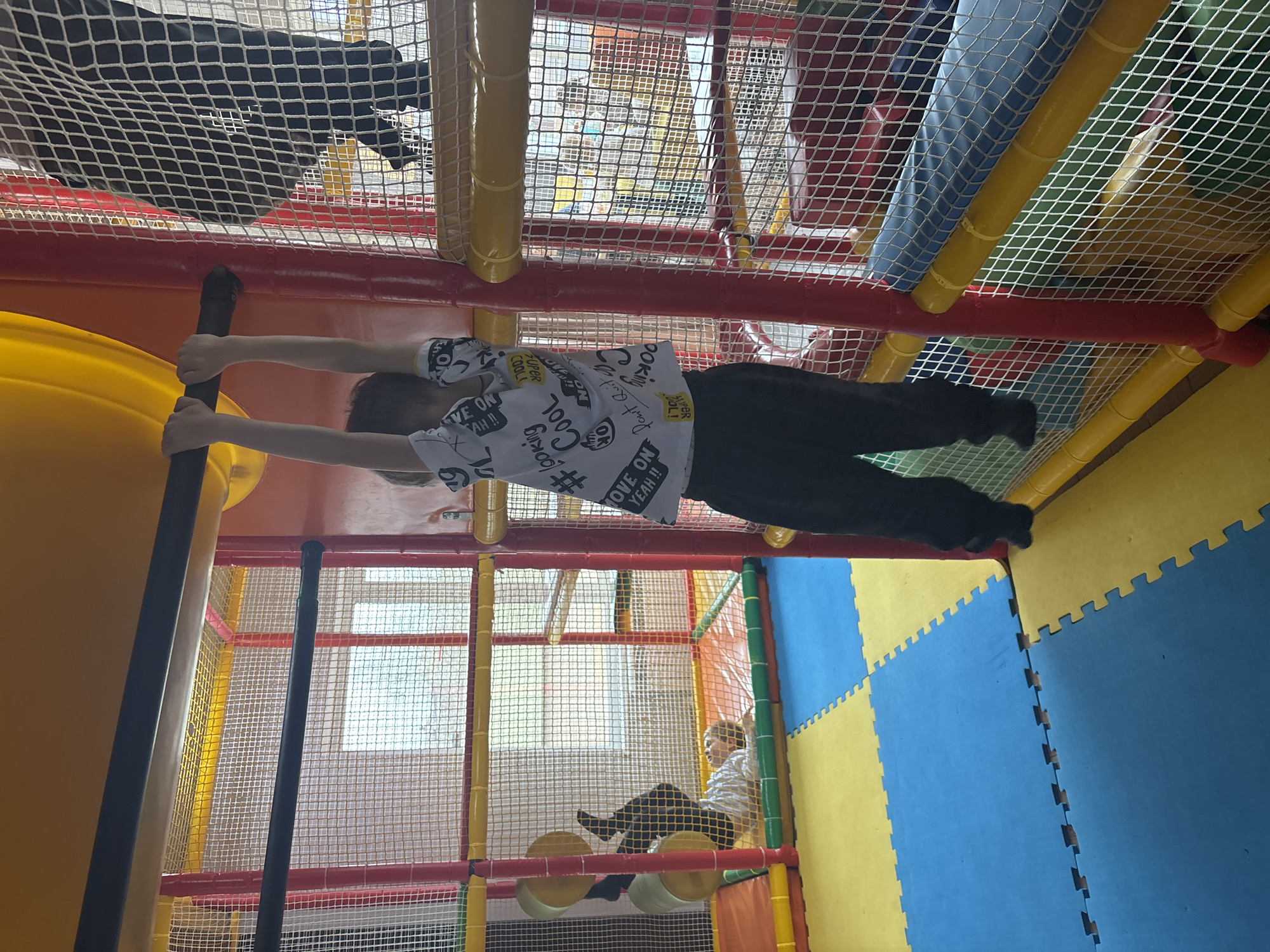 The image shows an indoor playground, likely in a family entertainment center. A young boy is hanging from a vertical pole within the play structure, extending his arms. The playground features colorful padding on the floor and structures with a network of netting and tunnels. Another child is visible in the background sitting on a padded structure within the play area. Based on the tags in the image metadata, this photo was taken in the city of Chaikovsky, Russia.