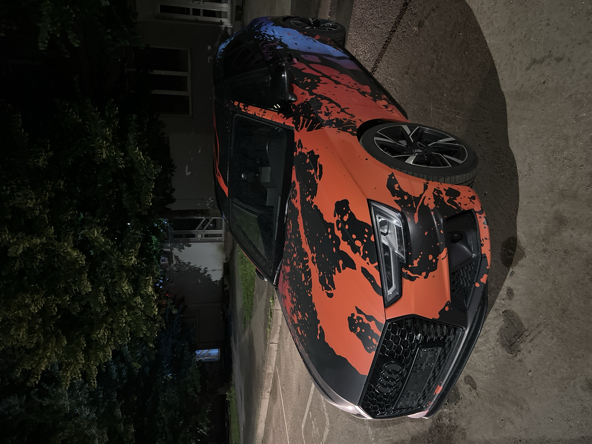 A black Audi A4 Avant with an orange and black abstract splatter graphic wrap is parked on asphalt in what appears to be a residential area at night. The car's front end is visible, showing the grille and headlights. The graphic wrap covers the hood, front fenders, and extends along the side of the car, transitioning to blue on the rear quarter panel. A large tree with dark foliage hangs over the car from the left. In the background, a building with windows is visible, and a streetlamp casts light on the scene. The ground is a mix of asphalt and dirt, with some stains or marks present. The overall lighting suggests it is nighttime, possibly with some ambient street lighting. This image was taken in Krasnodar, Russia.