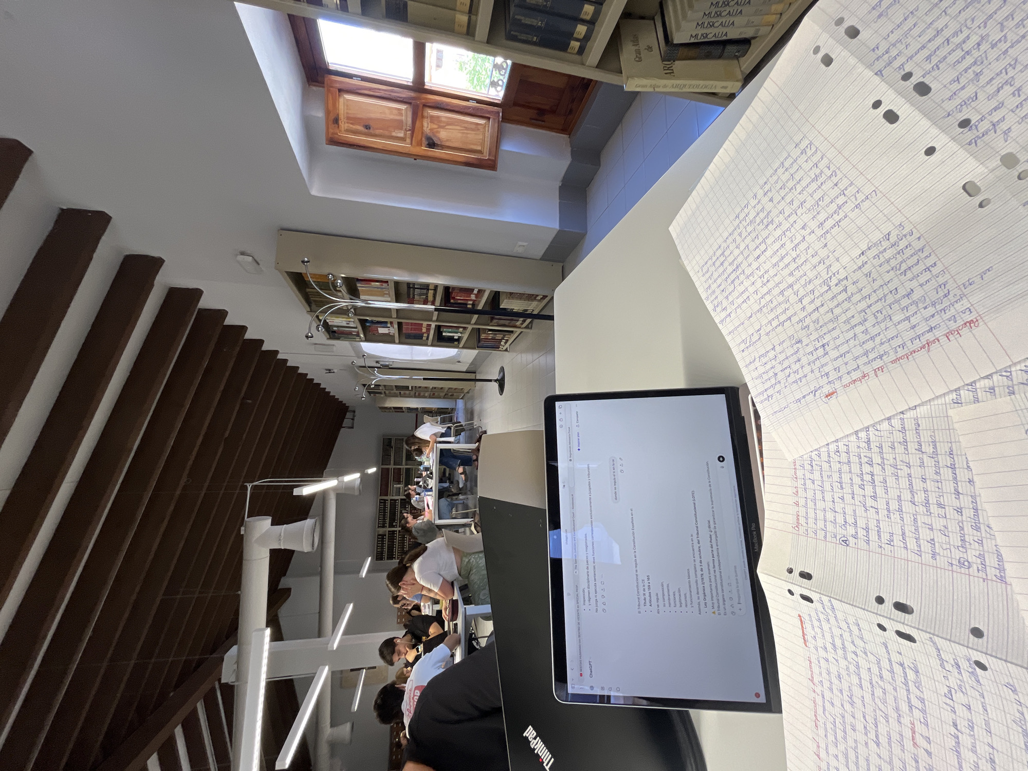 The image shows a well-lit study area in a library in Torrent, Spain. The view is from a student's perspective at a table with notes and a laptop. Several other students are studying at the tables. Bookshelves line the walls, and there are skylights.