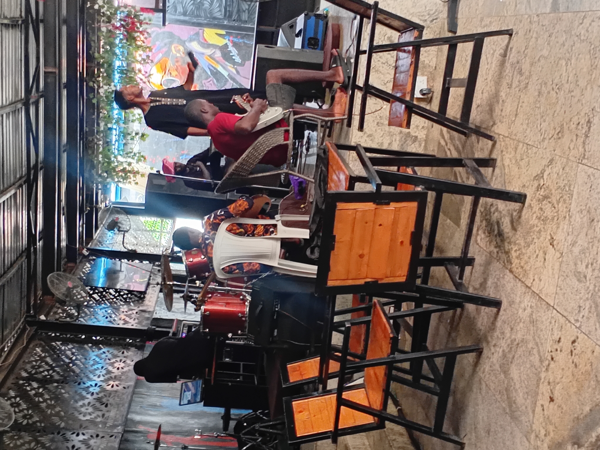 The image depicts an indoor music performance scene, likely at a venue in Howayi, Nigeria. In the foreground, several wooden chairs with black metal frames are arranged on a tiled floor, some facing a stage area. The stage itself features a drum set, musical equipment, and several individuals.
A person in a black outfit is seated at a drum kit, facing away from the camera, with a laptop in front of them. Another person, wearing a red shirt and dark shorts, is seated on a woven chair and actively playing a white electric guitar. Behind them, other individuals are visible, including one in a black garment and another with a pink head covering. A large, colorful mural or artwork is displayed on the wall behind the stage. The environment appears to be an open-air or partially covered space, with a corrugated metal roof structure visible above. Decorative greenery hangs from the ceiling. The lighting suggests it is either late afternoon or evening, with ambient light from the venue and possibly some stage lighting. The overall atmosphere appears to be casual and focused on the musical performance.