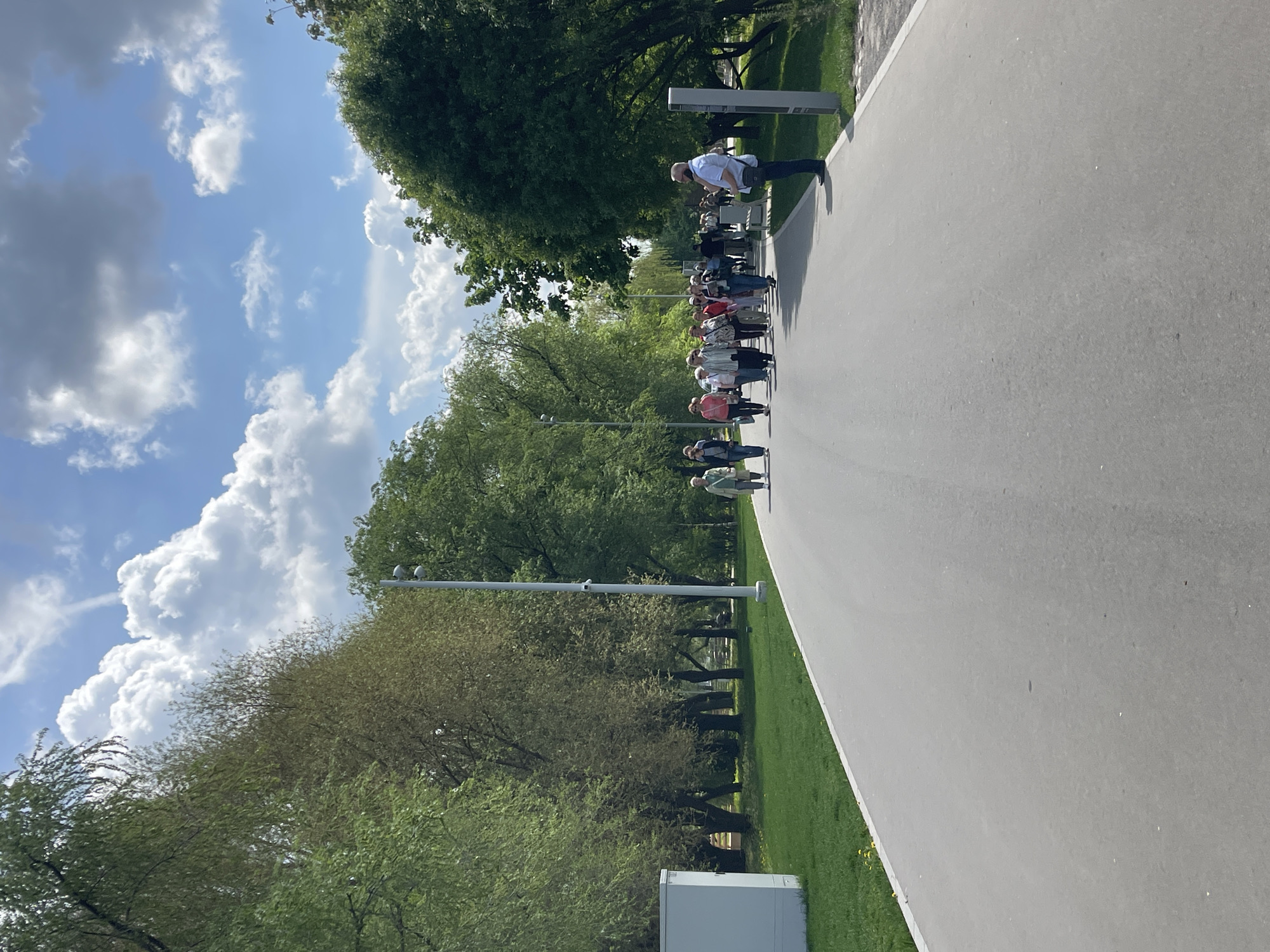 The image shows a paved walkway in Moscow, Russia, lined with trees and grass. A line of people, mostly older adults, are walking along the path, presumably in a group. A man in a white shirt stands on the path, appearing to be observing or guiding the group. The sky is blue with fluffy white clouds. Streetlights are visible along the path.