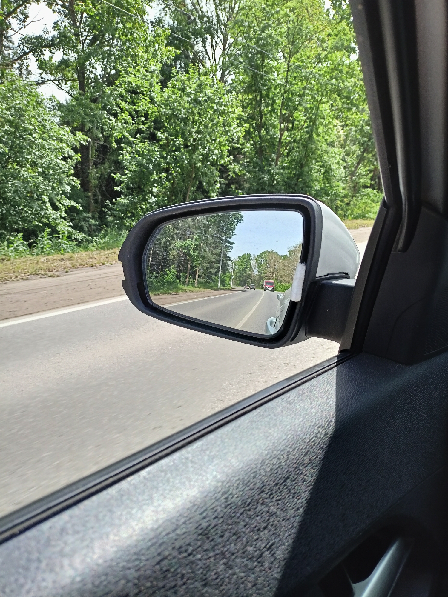 The image, taken from the interior of a vehicle, captures a reflection in the side mirror. The reflection shows a road with trees lining both sides and a red truck driving in the distance. The foreground of the image features the car's interior, including the door panel and the side mirror housing. The scene appears to be a rural or suburban area, possibly on a highway, given the presence of a road and natural landscape. The overall impression is one of travel through a verdant environment on a clear day. The specific location in Chaikovsky, Russia cannot be determined from the visual information provided.