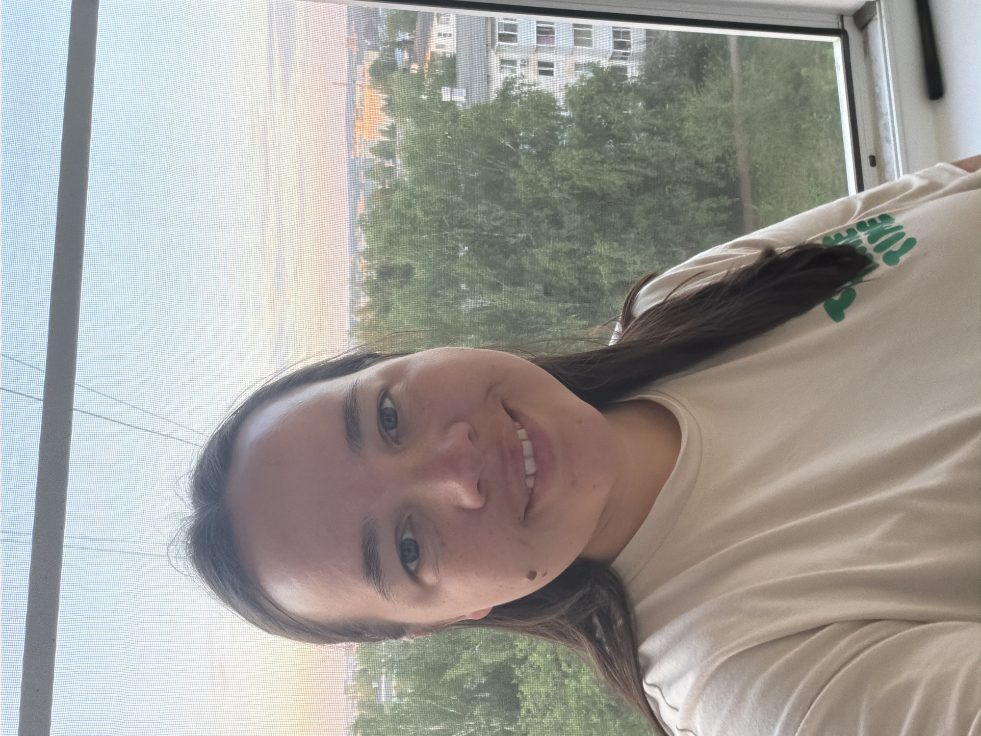 A young woman poses for a selfie indoors near a window overlooking the city of Chaikovsky, Russia. The background shows a building in the distance behind a line of trees. The foreground is dominated by the woman with long brown hair wearing a light-colored t-shirt with green accents. The window has a screen. The time appears to be daytime.