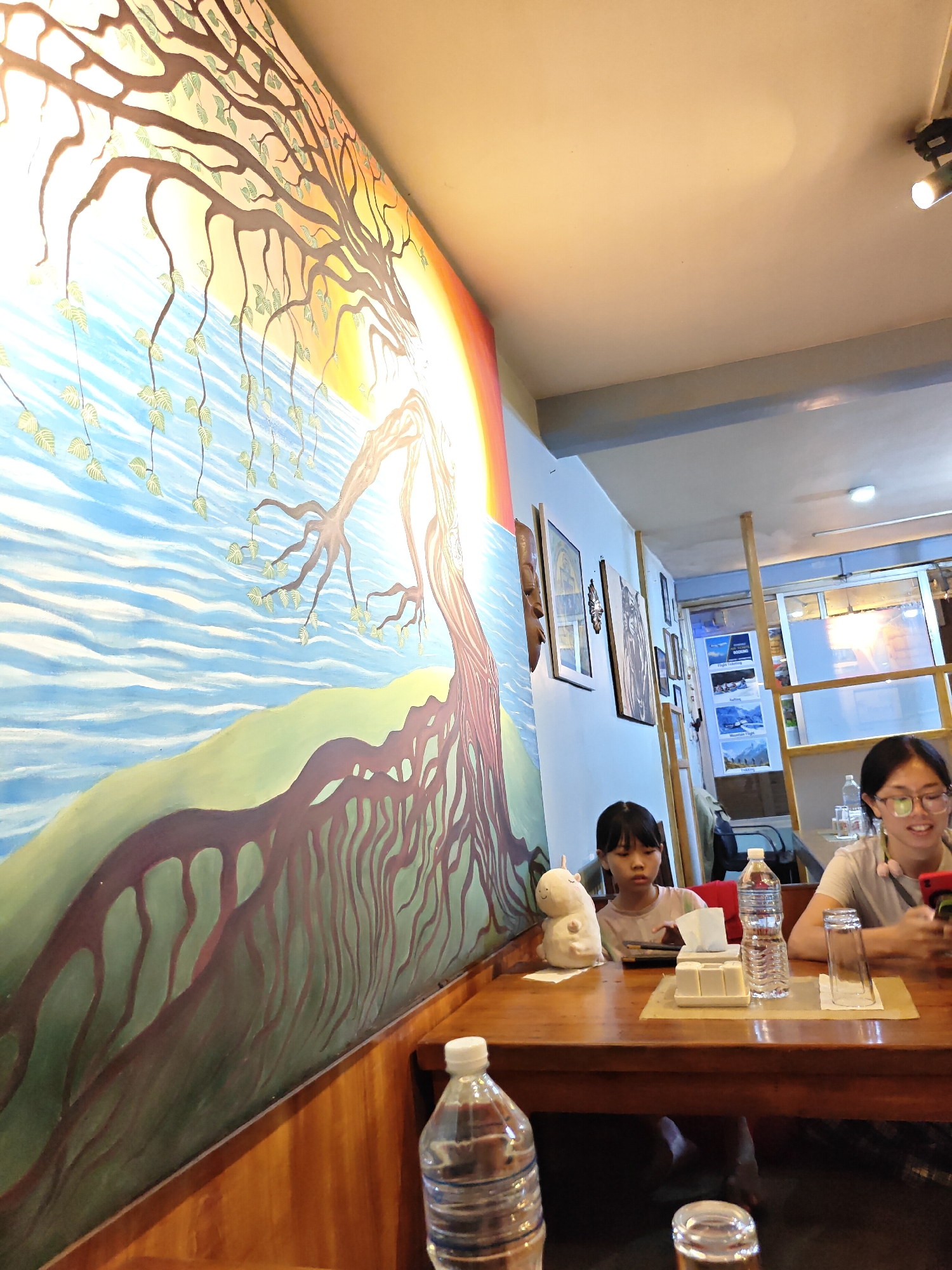 The image depicts the interior of a restaurant in Kathmandu, Nepal. The left side of the frame is dominated by a large, vibrant mural on the wall, featuring a stylized tree with flowing branches and roots against a backdrop of blue water and a bright yellow and orange sun. The mural extends across the wall, creating a colorful and artistic ambiance.
In the foreground, a wooden table is set with various items. Two young girls are seated at the table. The girl on the left, with dark hair, is looking down, seemingly engaged with something on the table. A white stuffed animal, resembling a unicorn or llama, is placed next to her. On the table, there are also tissue dispensers, a bottle of water, and a glass.
The girl on the right, wearing glasses and a light-colored t-shirt, is looking towards her right, holding a mobile phone in her hand and appearing to be using it. Another bottle of water and a glass are visible in front of her.
The background shows more of the restaurant's interior, with wooden pillars, framed pictures on the wall, and a glass-partitioned area with what appears to be stairs or an escalator visible through it. The lighting in the restaurant is warm, with spotlights visible on the ceiling. The overall impression is of a casual dining setting with artistic decorations.