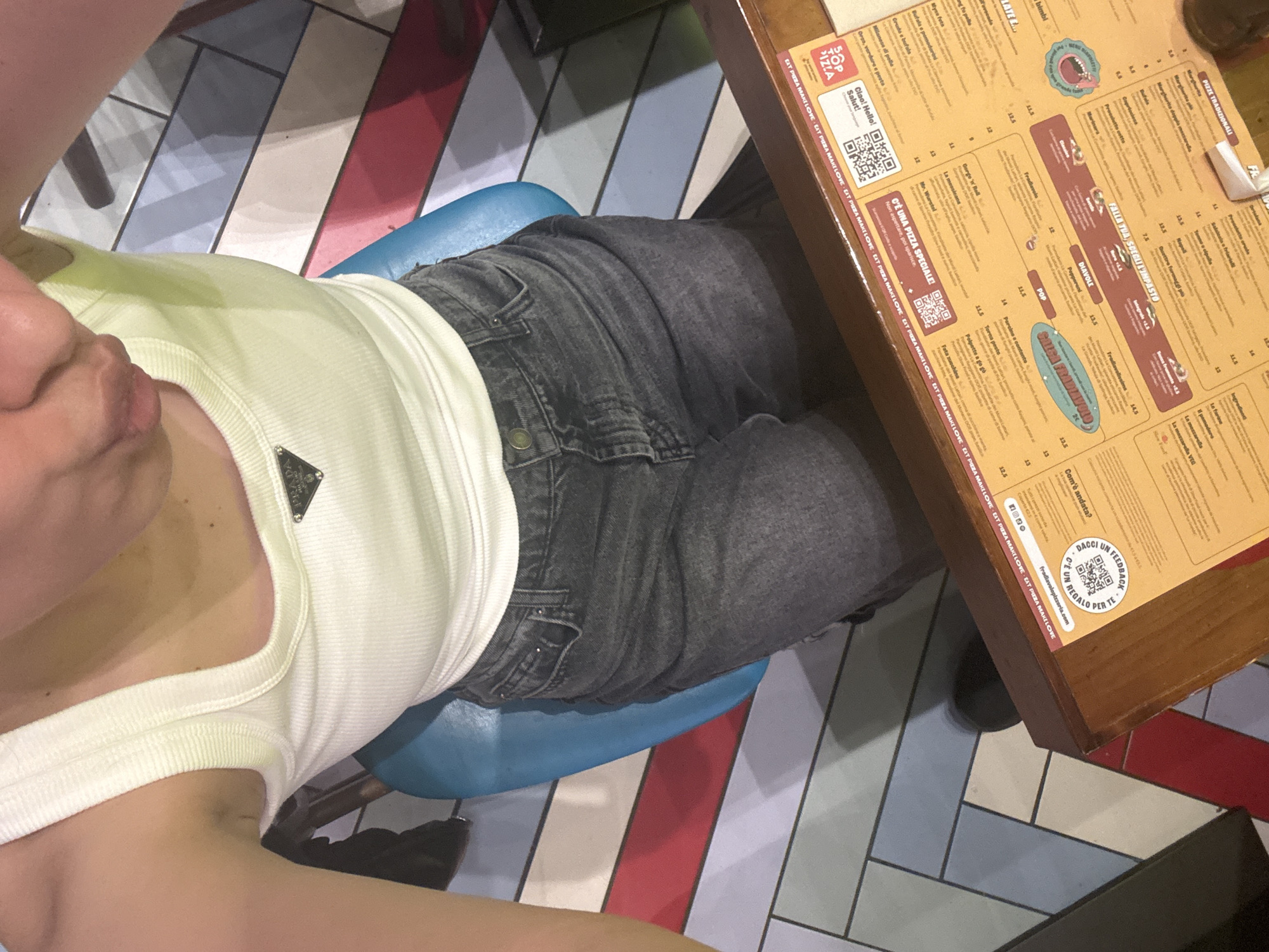 The image depicts a person seated at a table in what appears to be a pizza restaurant. The person is wearing a white tank top and jeans. On the table is a menu from a restaurant named "Fradiavolo". The menu lists various pizza options. The table itself is wooden and the floor is tiled in a herringbone pattern with red, white, blue, and gray tiles. Based on the menu being in Italian, and the restaurant name, it is possible this is located in Milan, Italy.