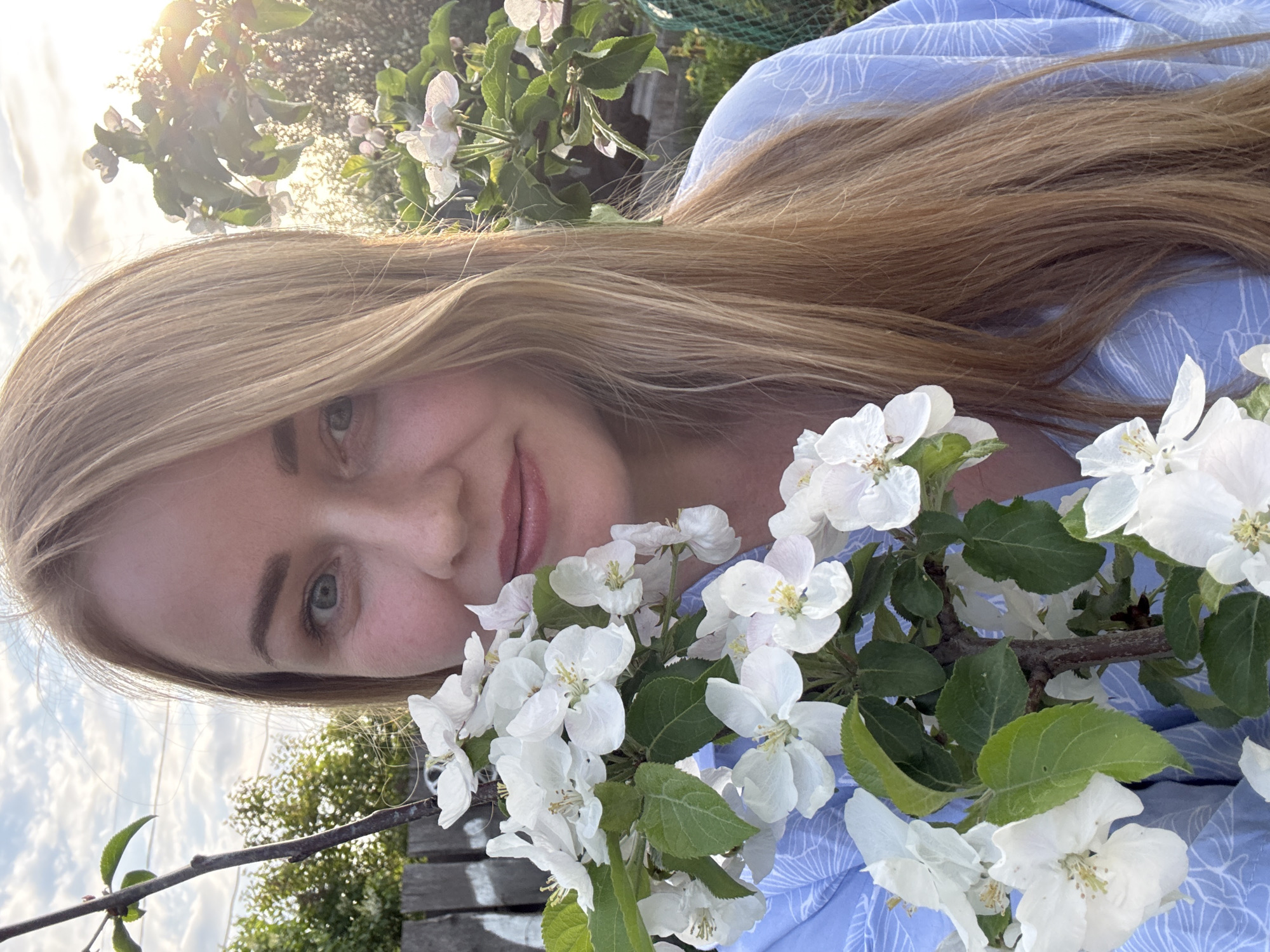 A young woman with long blonde hair is holding a branch of white apple blossoms, smiling slightly. She is wearing a light blue patterned shirt. In the background are green trees and branches, with sunlight filtering through. The location is likely outdoors in the city of Chaikovsky, Russia, given the metadata.