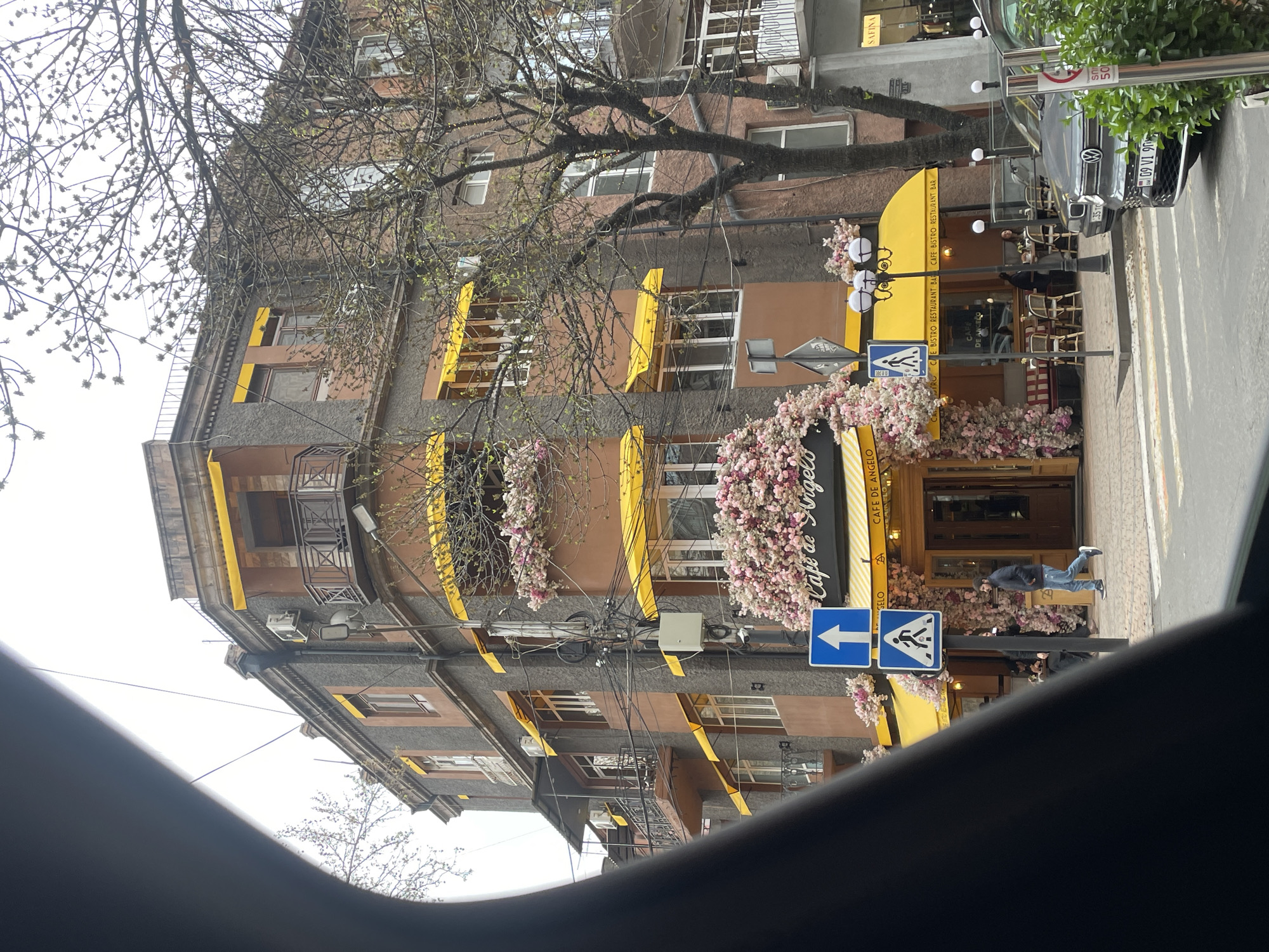 The image depicts the exterior of "Cafe de Angelo" in Yerevan, Armenia. The building is multi-story with a mix of brown and gray textured facades, windows adorned with yellow awnings, and floral arrangements cascading around the cafe's entrance. A street sign with a left-pointing arrow and pedestrian crossing symbol is visible. A man in a dark jacket and blue jeans is walking past the cafe. A gray Volkswagen is parked nearby, bearing license plate "35 TO 09 VI." The scene is observed from within a vehicle, as part of the dashboard is visible at the bottom of the frame. Bare tree branches are visible above the building.