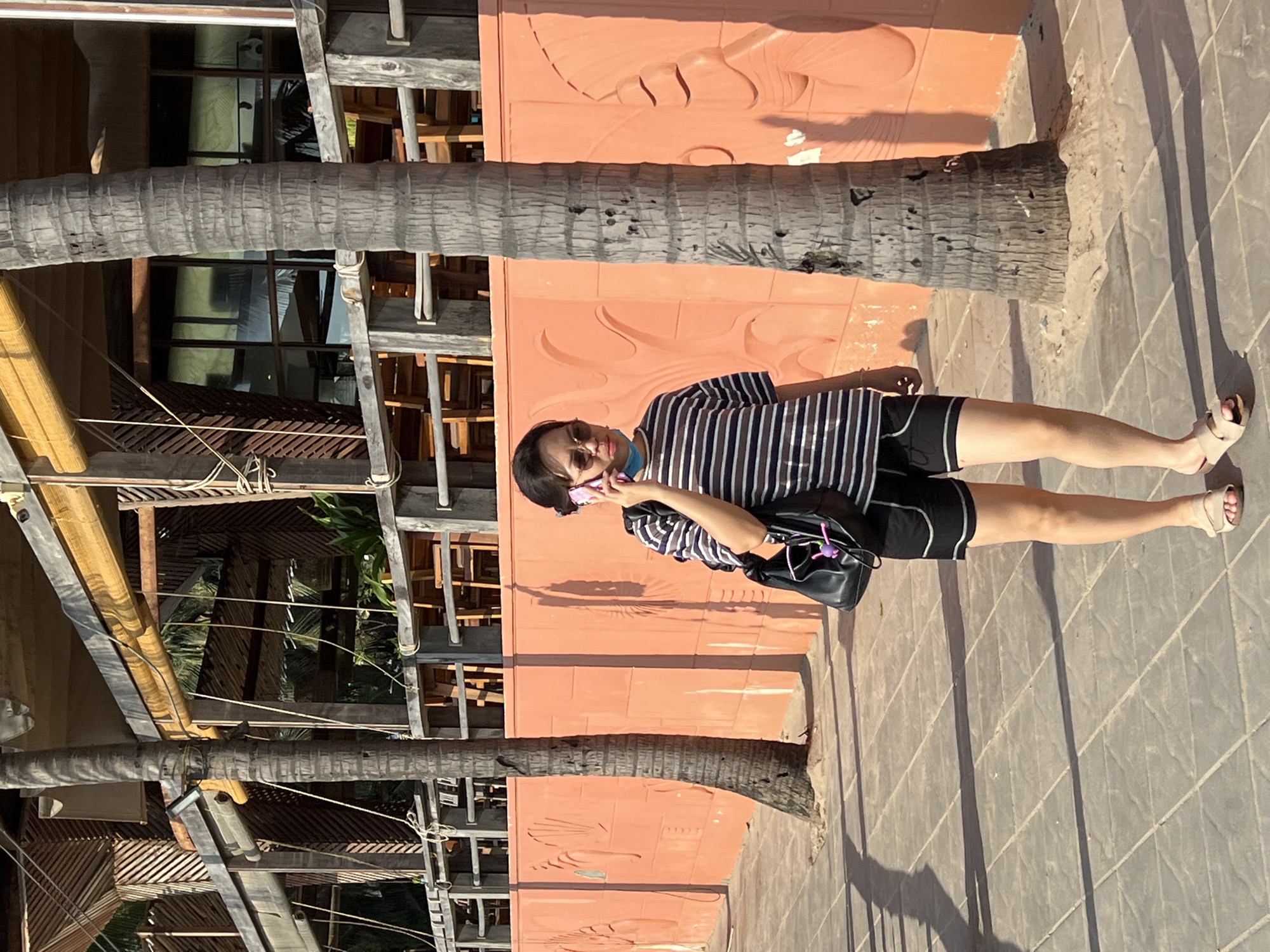 A woman stands outdoors, holding a mobile phone to her left ear. She wears a black and white striped shirt, black shorts, and tan sandals. A black purse hangs from her left shoulder. The woman stands in front of a peach-colored wall with carved details and a tree trunk. In the background, there is a building with weathered wooden structures. The ground is paved with gray tiles. The location is Nha Trang, Vietnam.