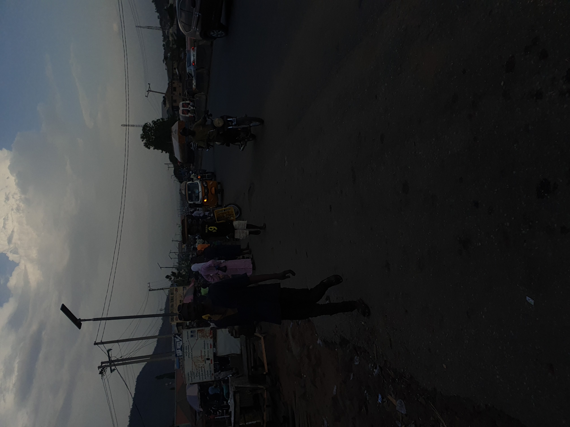 The image depicts a street scene in Lokoja, Nigeria. A group of people is gathered near a yellow tricycle taxi (keke). Individuals are walking along the road. A person is prominently positioned in the foreground, seemingly in motion. There's a motorcycle parked further down the road. Buildings are visible on either side of the street, along with utility poles and wires overhead. The sky is cloudy. The lighting suggests it's either early morning or late afternoon.
