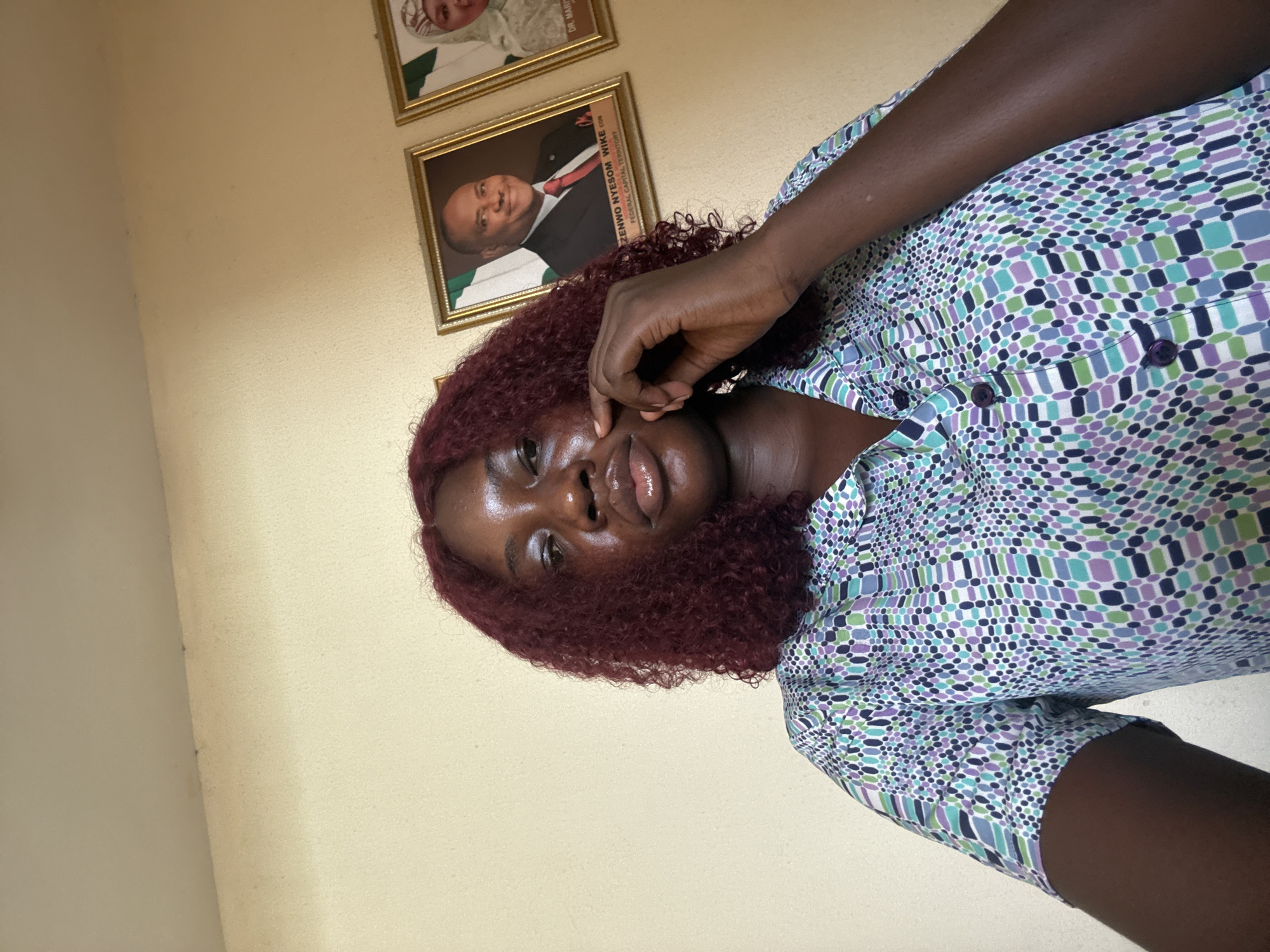 A medium-shot photograph depicts a dark-skinned woman with burgundy curly hair, wearing a patterned shirt with blue, purple, and white geometric designs. She is touching her face with her right hand, and looking at the camera with a slight smile. Behind her, on a light yellow wall, are two framed portraits, one of a man in a suit and the other not easily discernible. The name "ZENWO NYESOM WIKE" and "FEDERAL CAPITAL TERRITORY" are partly visible on the frame of the portrait, which indicates a location in Abuja, Nigeria.
