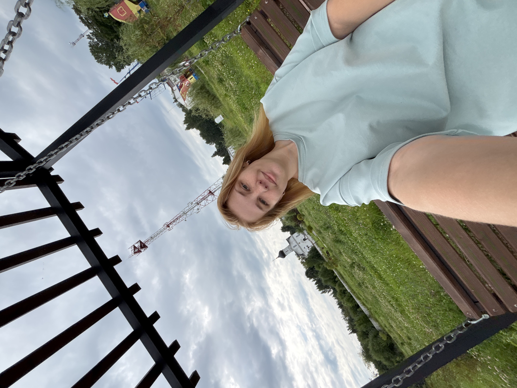 A young woman with blonde hair takes a selfie while sitting on a wooden swing in what appears to be a park or outdoor recreational area. She is wearing a light blue t-shirt. In the background, a white church with a dark dome is visible, surrounded by green trees and a field of grass with small white flowers. A radio tower is also present in the distance, along with other structures that suggest a town or amusement park. The sky is overcast. This scene is located in the city of Breytovo, Russia.