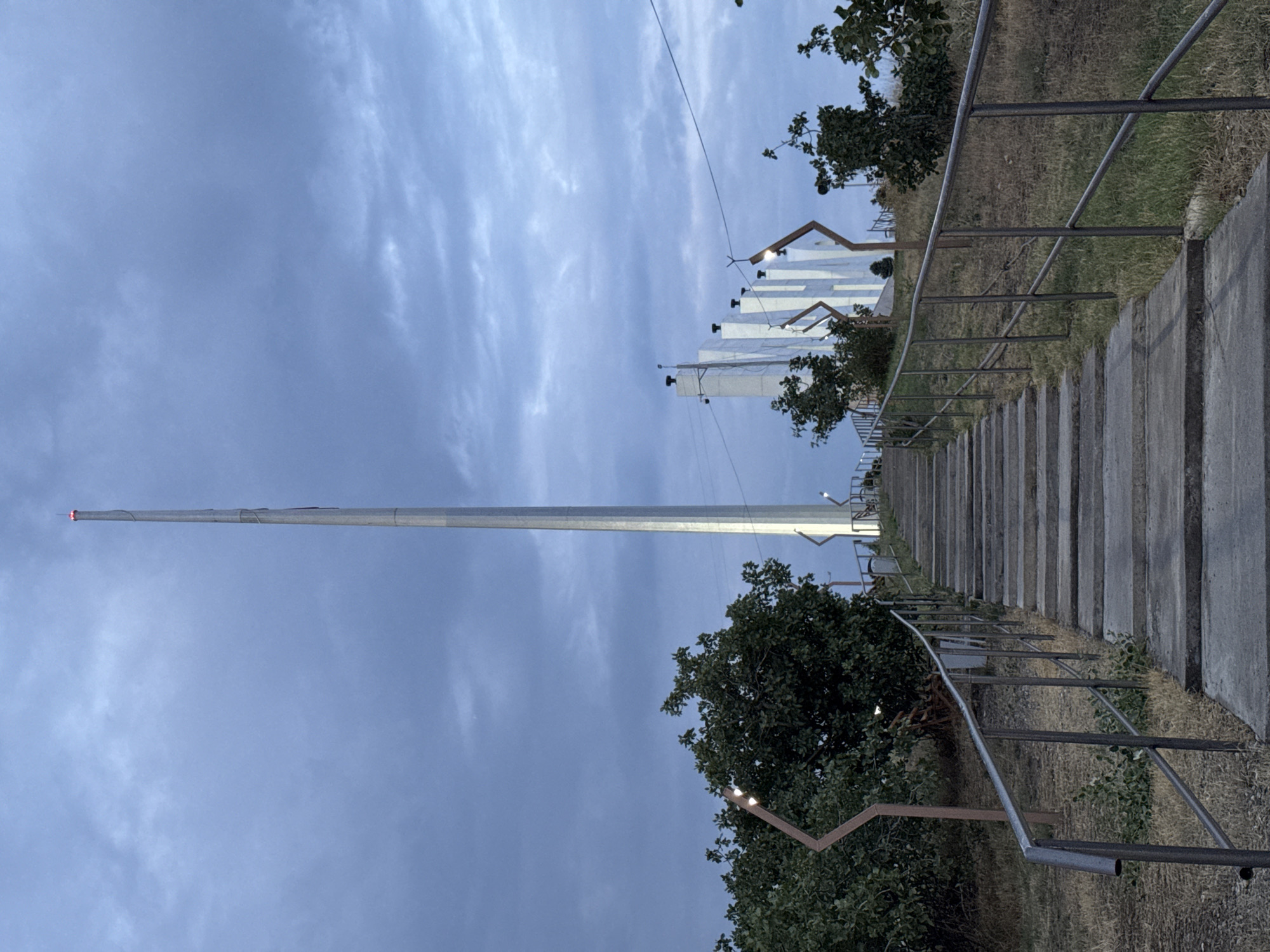 The image depicts a tall, slender metallic flagpole topped with red lights, situated in the city of Manas, Kyrgyzstan. A wide concrete staircase with metal railings leads upwards towards the flagpole. The surroundings are characterized by dry grass, sparse trees, and modern-looking white structures in the background. The sky is overcast with a palette of blue and grey clouds, indicating either dawn or dusk. There are no people visible in the scene.