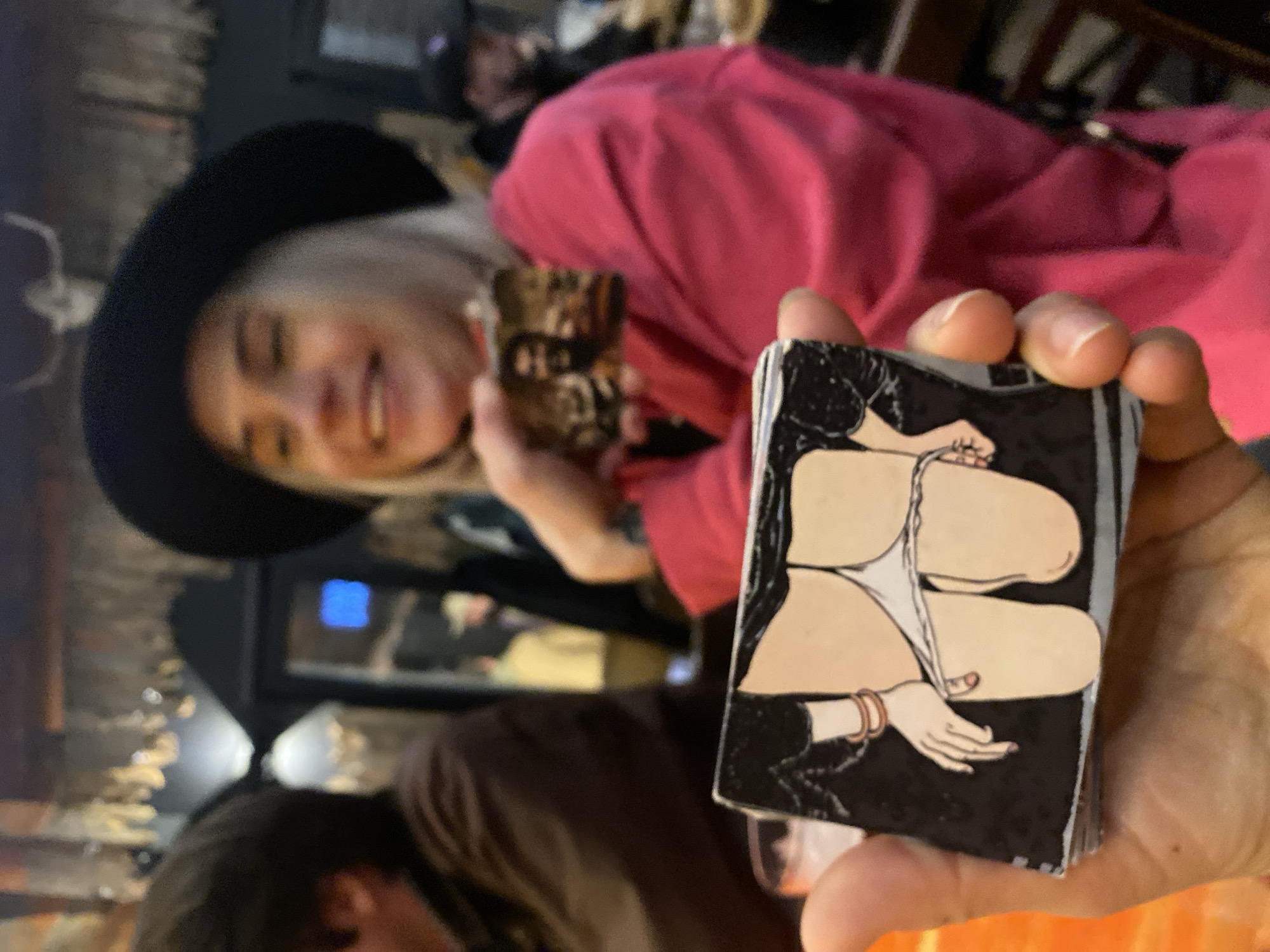 The image shows a person holding a deck of cards with an illustration of a partially nude female figure in front of a slightly blurred background. The background shows a person wearing a black hat and a person wearing a pink top and holding a small decorative item. Another person is partially visible on the bottom left. The environment suggests an indoor setting, possibly a bar or restaurant, with wooden chairs visible in the background. There is no information to determine the location is Moscow, Russia.