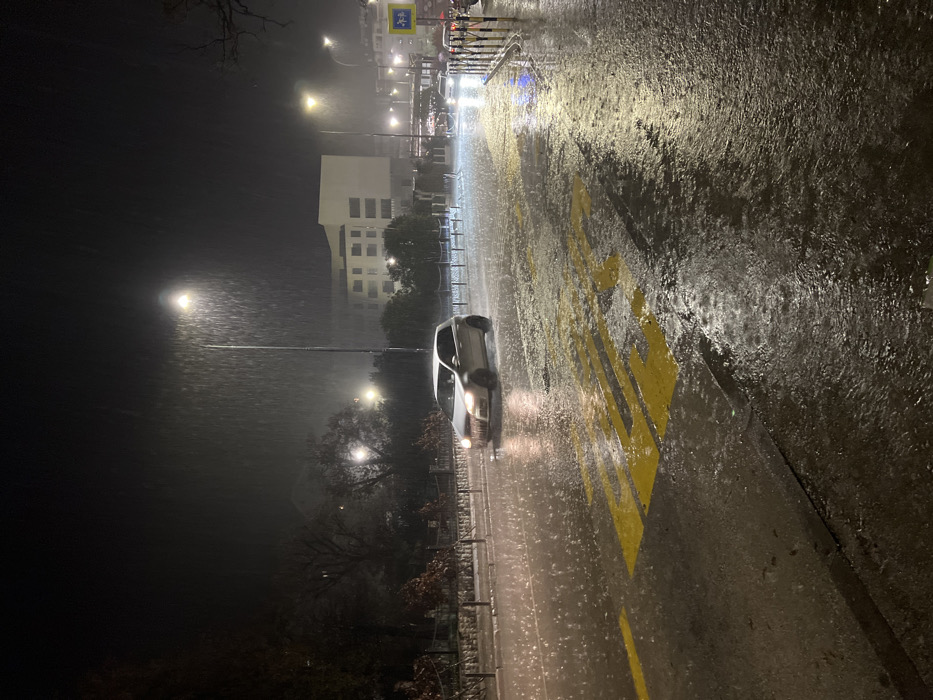 Rainy night street scene featuring a single car with headlights on, driving on a wet road with yellow markings. A tall, light-colored building is visible in the background, partially illuminated by streetlights. The rain is dense, creating a blurred effect and reflecting light from street lamps and vehicle lights. The overall atmosphere is dark and wet.