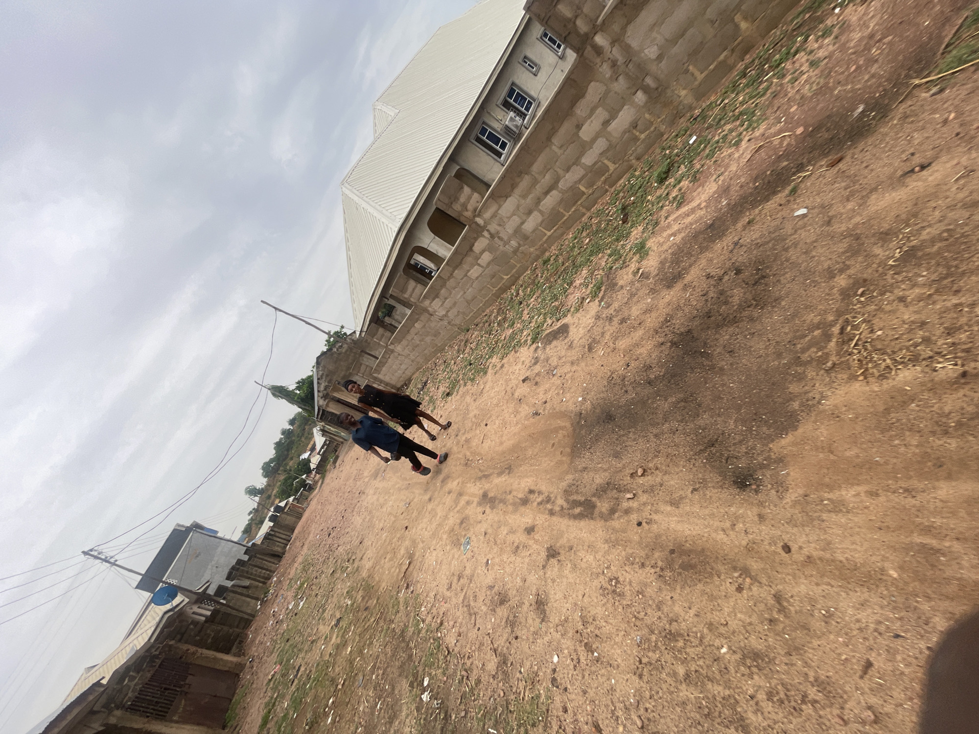 Two people, a male and a female, are walking on a dirt road. The male is wearing a blue shirt and black pants. The female is wearing a black dress. They are walking away from the camera. To their right is a building with a white roof and brick walls. The road is dry and dusty. In the background, there are other buildings and trees. The sky is overcast. The location is likely Lokoja, Nigeria, inferred from the prevalence of similar architectural styles in the region and the red soil.
