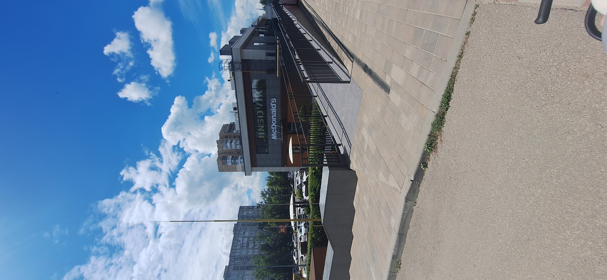 The image shows a modern McDonald's restaurant with a ramp leading to its entrance. The building is located in Bila Tserkva, Ukraine, as indicated by the presence of the McDonald's signage. The sky is a vibrant blue with scattered white clouds. In the background, there are apartment buildings and trees. The foreground features a sidewalk and a paved road. Cars are parked in a lot next to the McDonald's. The overall scene depicts a typical urban environment on a clear day.