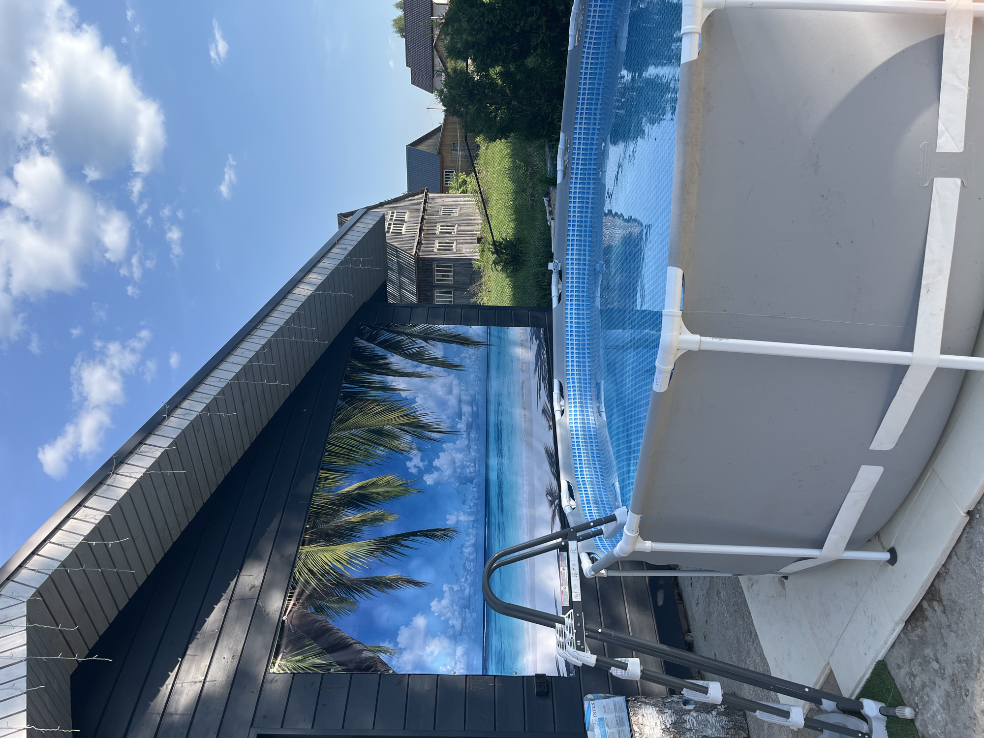 The image shows a backyard scene in Новая Чернушка, Russia, featuring a round, above-ground swimming pool. The pool is predominantly gray with white support structures. A ladder provides access to the water, which has a blue tile pattern lining. Adjacent to the pool is a dark wooden structure with a large tropical-themed print of palm trees and a beach scene. Beyond the pool and wooden structure, one can observe houses and greenery, indicating a residential area. The sky above is blue with scattered clouds. The overall environment suggests a summer day.