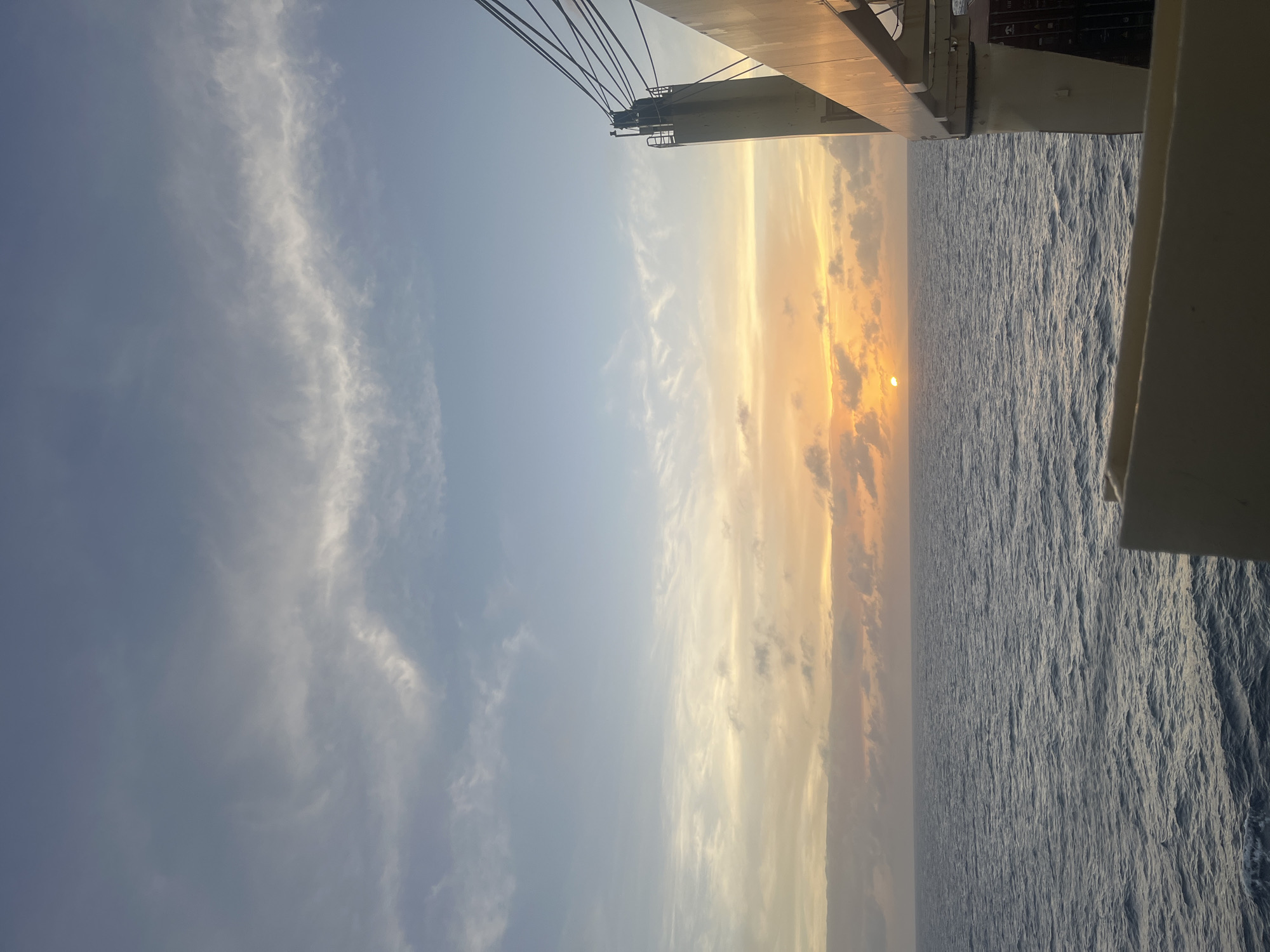 The image shows a sunset over the ocean, likely taken from a ship or boat. The sky is a gradient of blues and yellows, with the sun visible through a layer of clouds. The ocean is a dark blue with visible ripples and waves. Part of the ship's equipment is visible in the upper and right edges of the frame. The location is Unknown, Unknown.
