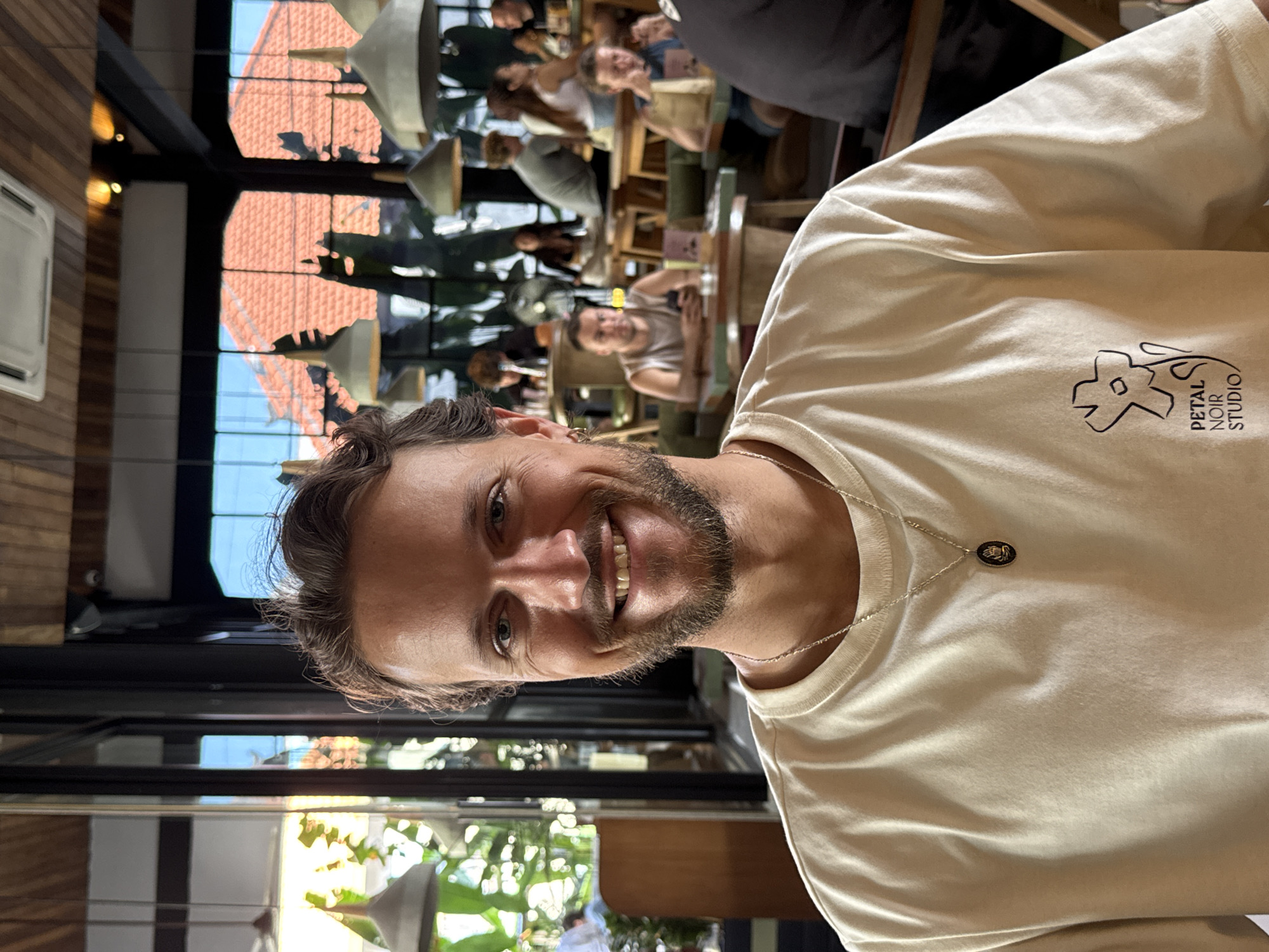 The image shows a man with short brown hair, a beard, and blue eyes wearing a light-colored t-shirt with "PETAL NOIR STUDIO" printed on it. He is also wearing a silver necklace with a dark-colored pendant. He is smiling and looking at the camera.
The background shows a restaurant or cafe setting with various people seated at tables. There is a view of the outside through the glass windows, showing trees and buildings, including roofs. The location is likely in North Kuta, Indonesia, as indicated in the instructions. The restaurant has hanging lamps and plants. There are several people in the background, some sitting at tables and others walking around. The setting suggests an outdoor or semi-outdoor dining area.